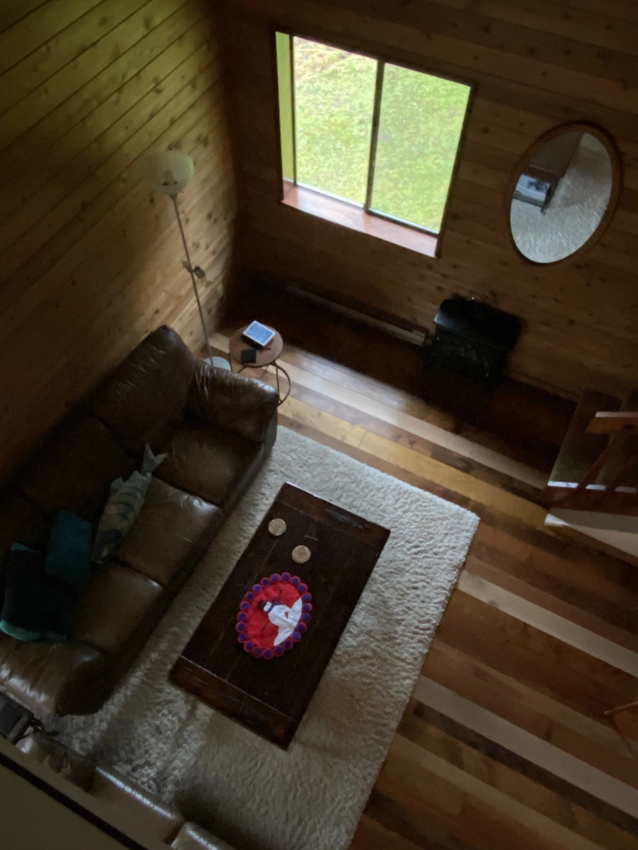 Living room with wooden walls and floors, a large window, a leather sofa with a blanket, a small side table with a tablet, a white rug, a coffee table with a Christmas-themed decoration, and a mirror on the wall.