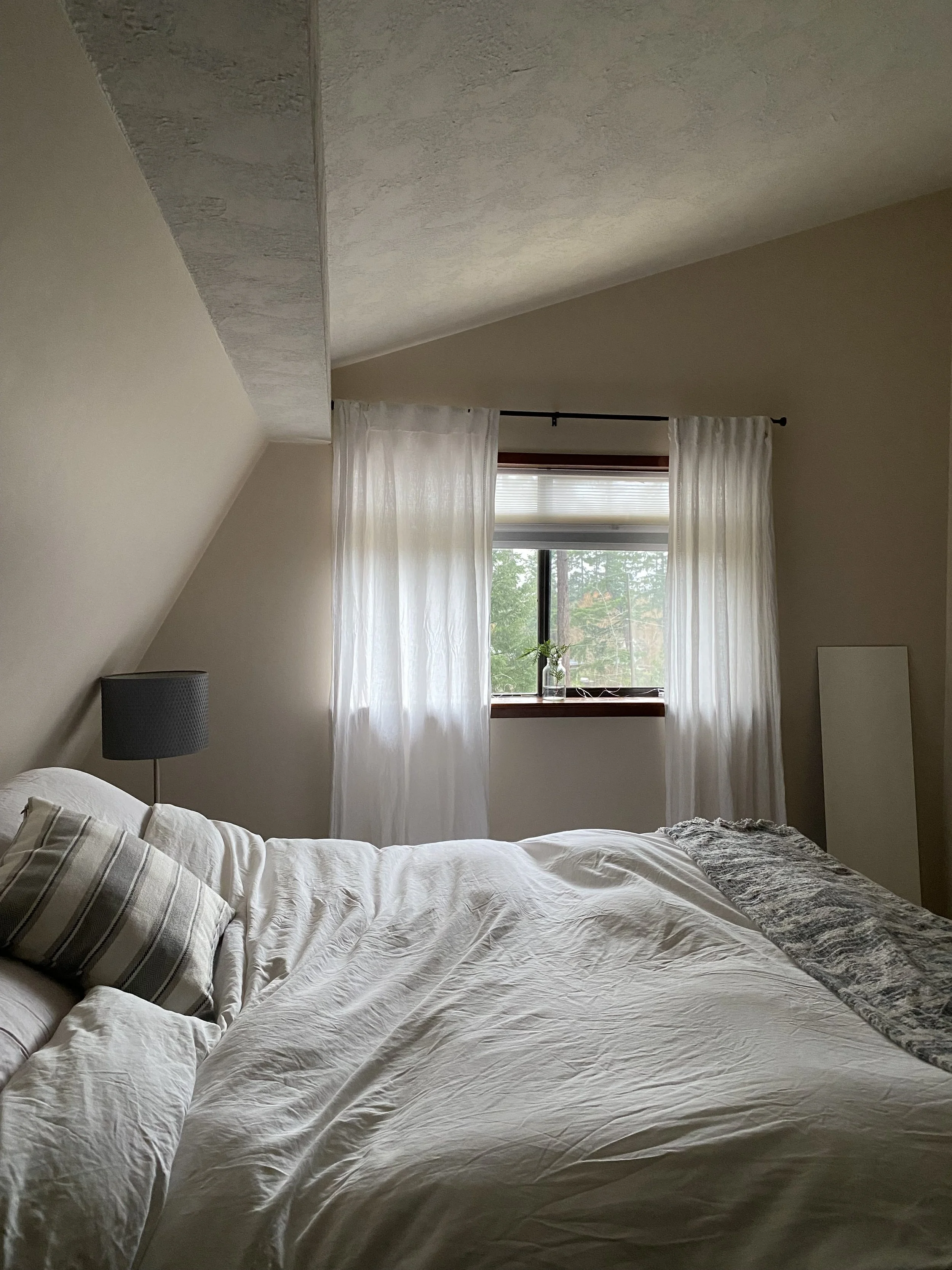 A cozy bedroom with a sloped ceiling, a bed with white bedding and striped pillows, a dark gray bedside lamp, and a window with sheer white curtains, allowing natural light to illuminate the room.