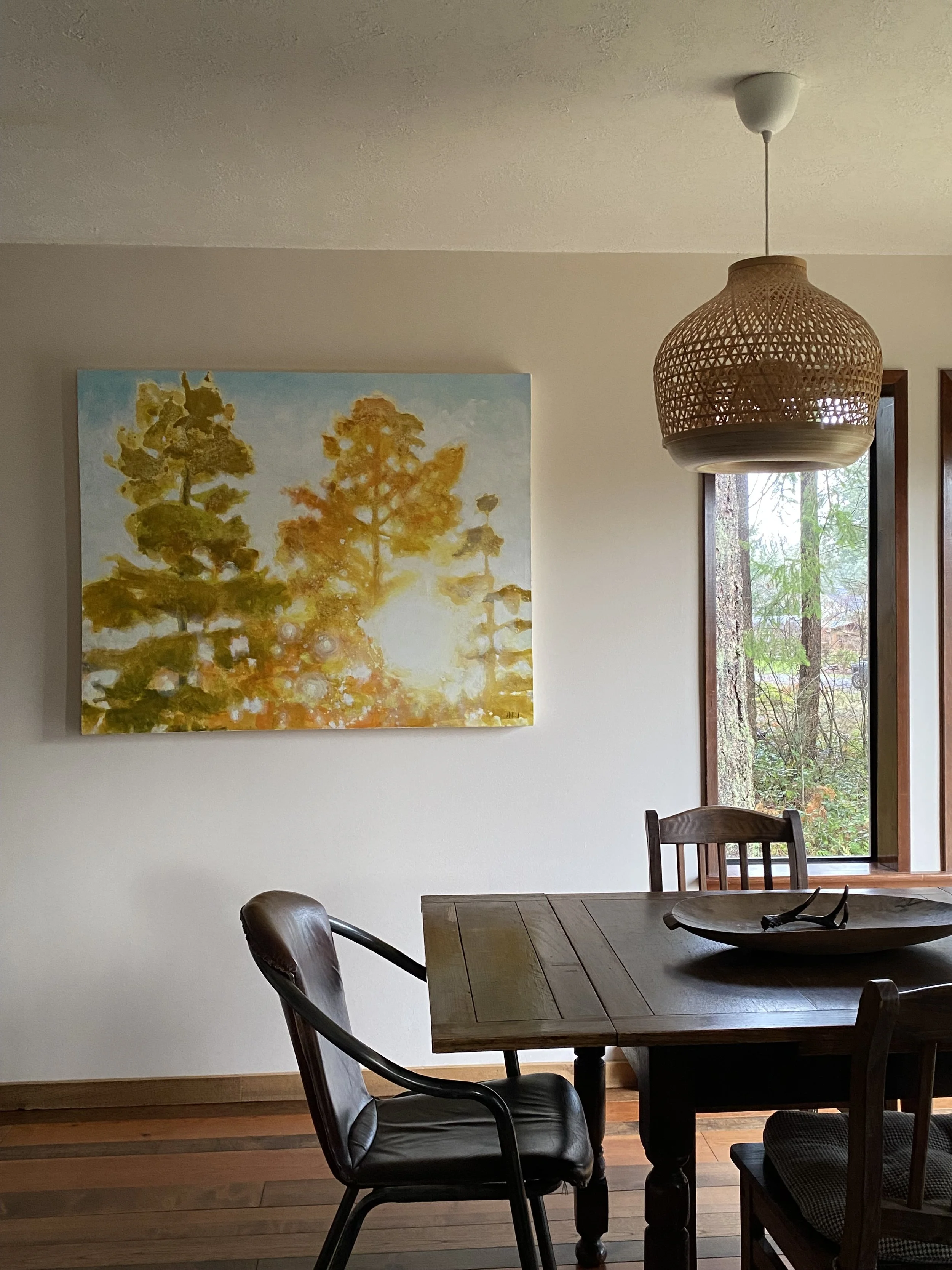 Comox Valley furnished short term rentals. Interior of a dining area with a wooden table, leather chair, wall art of trees with sunlight, a wicker pendant light, and large windows showing trees outside.