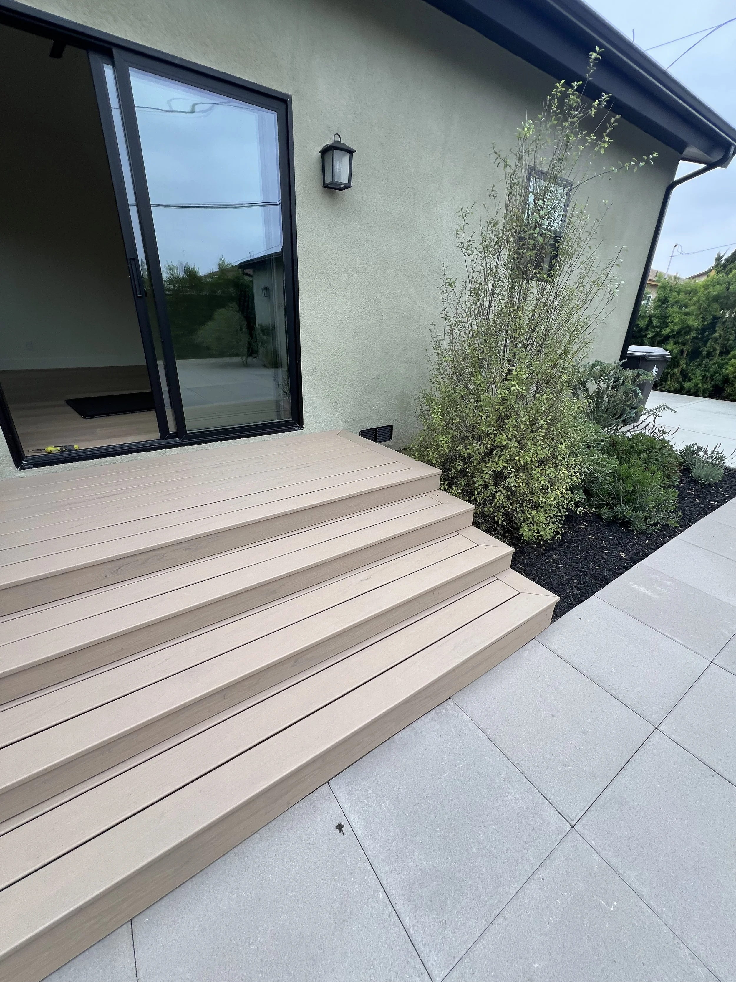 Back porch with beige wooden steps leading to a glass sliding door, a wall-mounted outdoor light, and a garden area with bushes and planting beds.