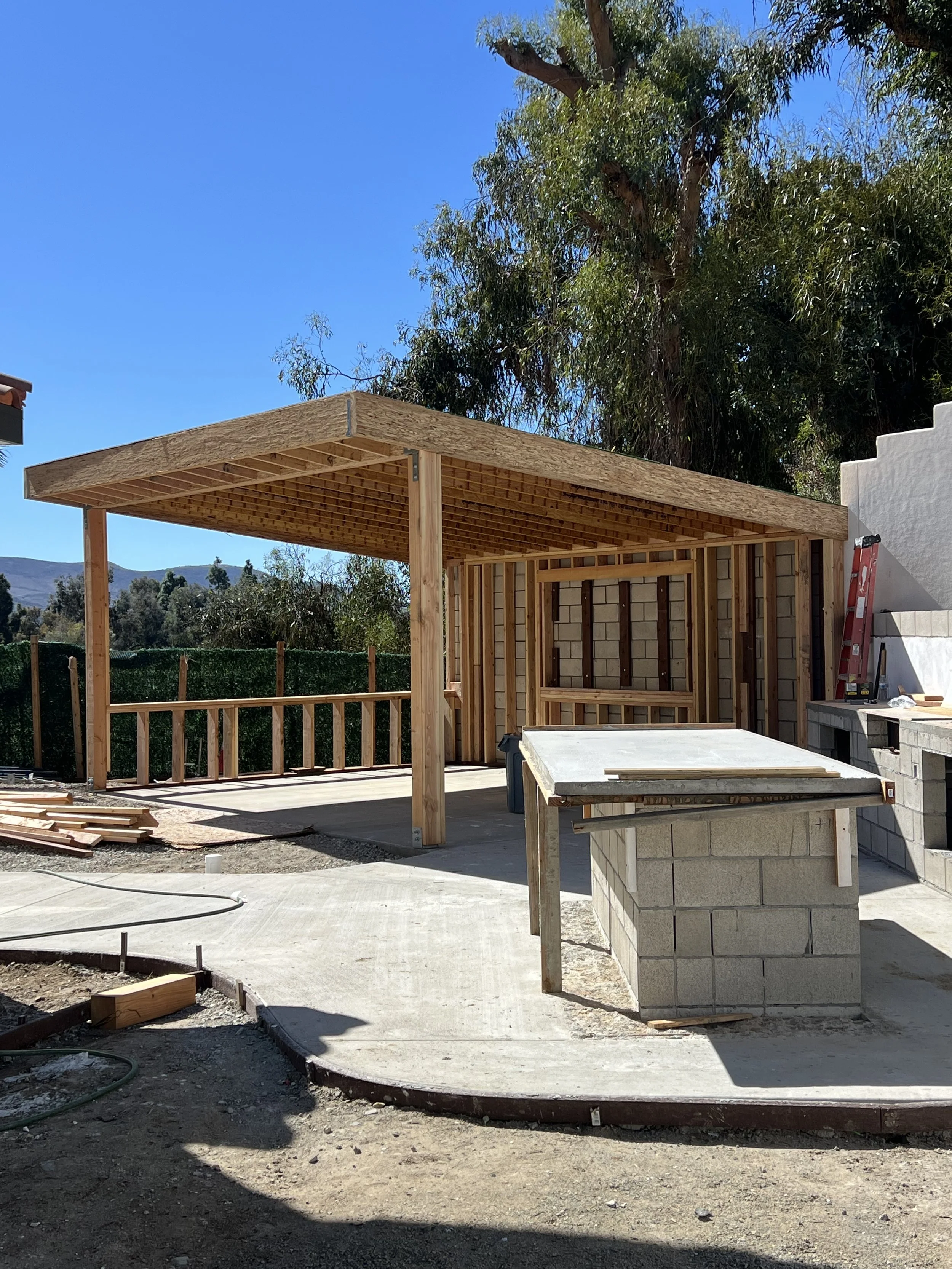 Construction site with a wooden shelter frame, concrete patio, and outdoor countertop under a clear blue sky.