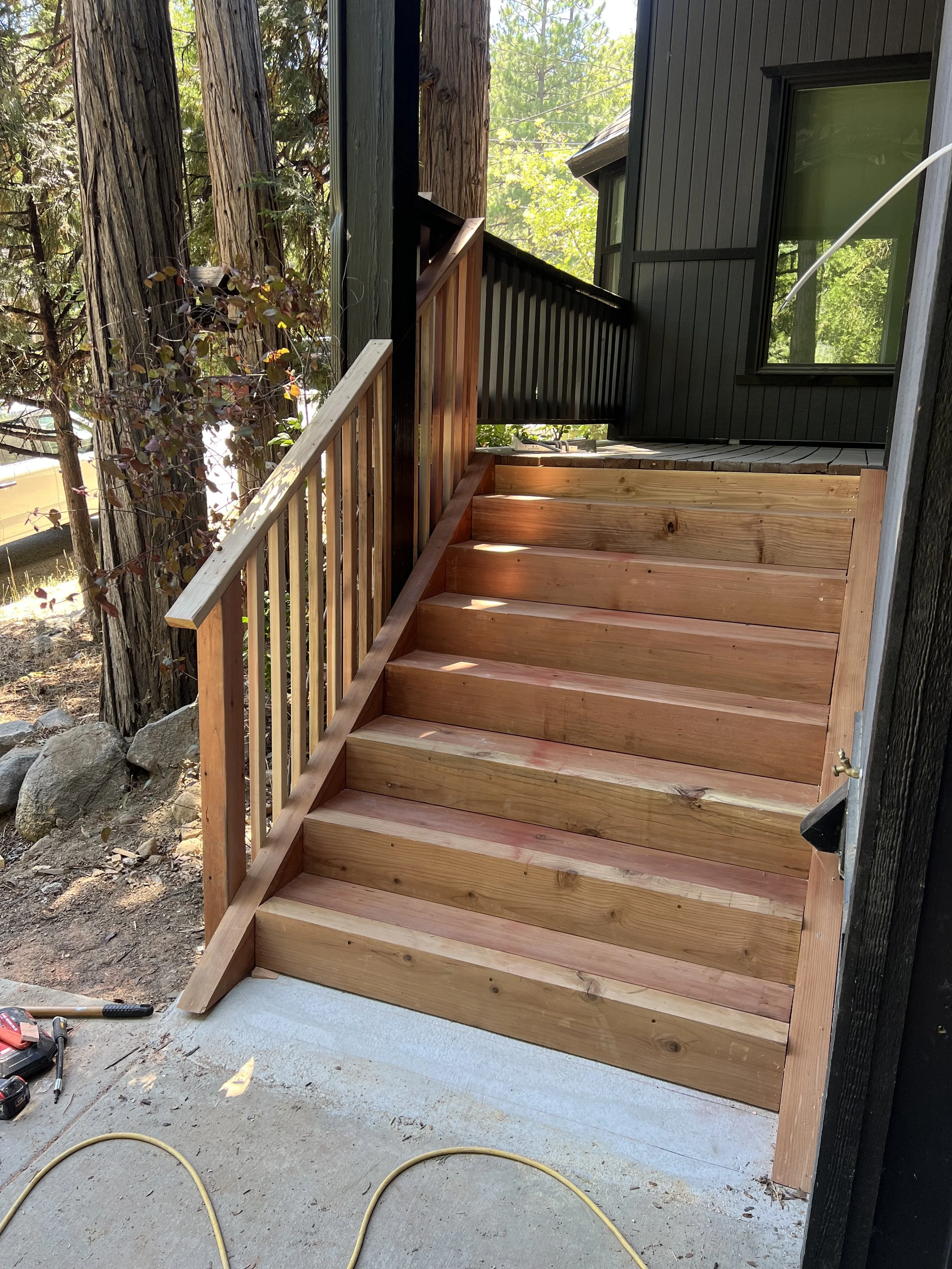 Wooden stairs leading up to a house with black exterior siding, surrounded by trees and natural landscape.