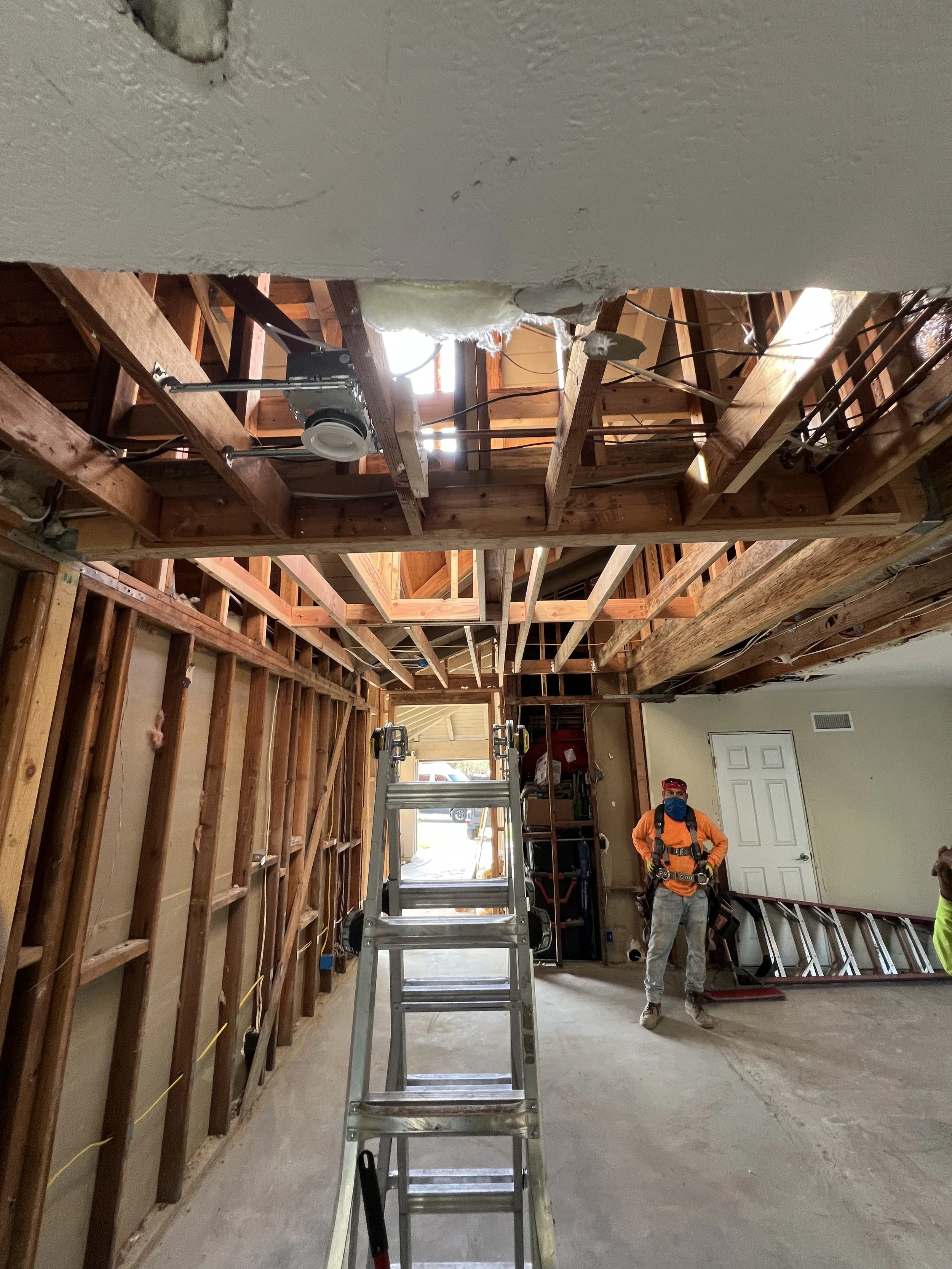 Construction workers in an unfinished room with exposed wooden beams and framing, a ladder, and construction tools.