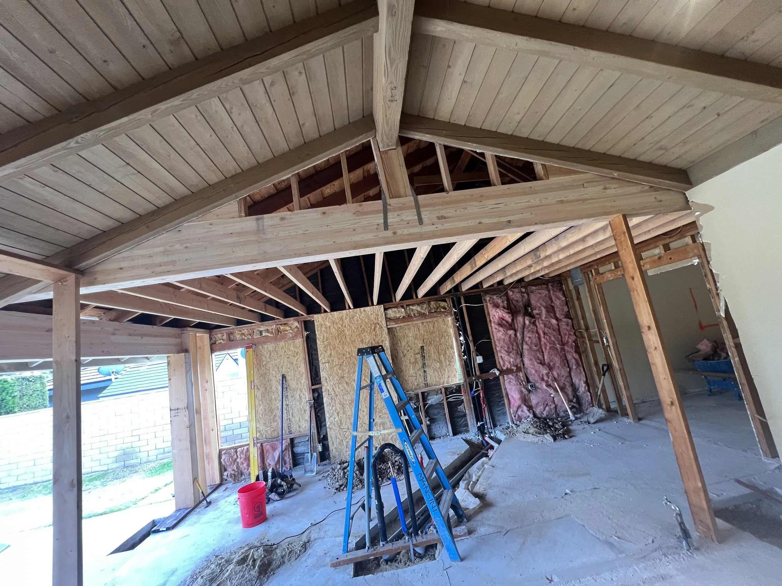 Interior view of a house under construction, showing exposed wooden framing, insulation, construction tools, and a ladder.