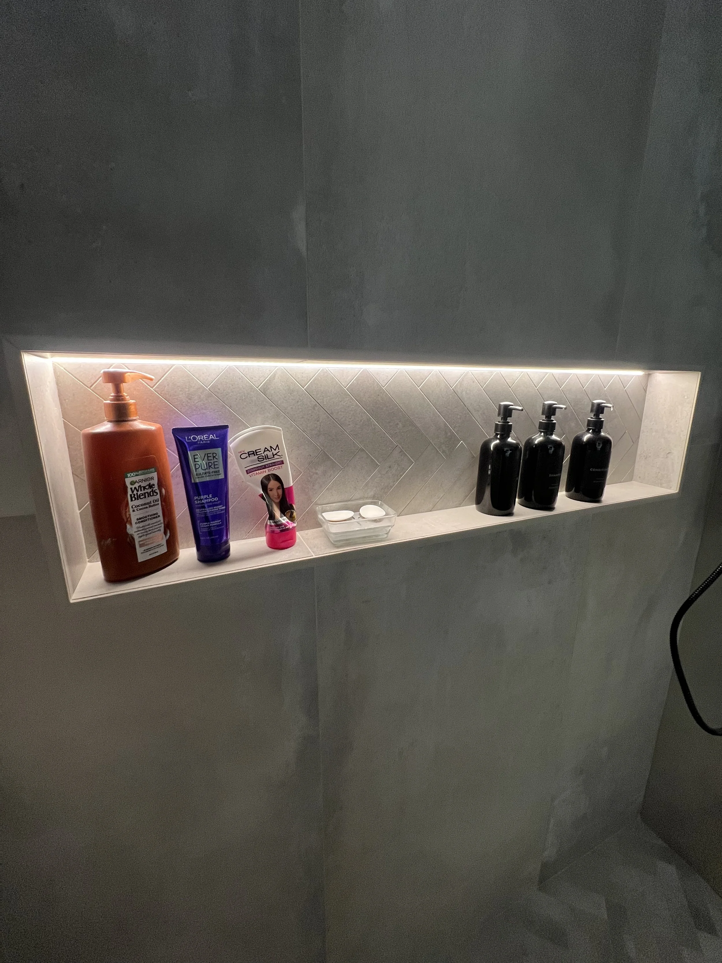 Recessed bathroom shelf with various personal care products, including shampoo, conditioner, body wash, face cleanser, soap, and lotion bottles, against a tiled wall with LED lighting.