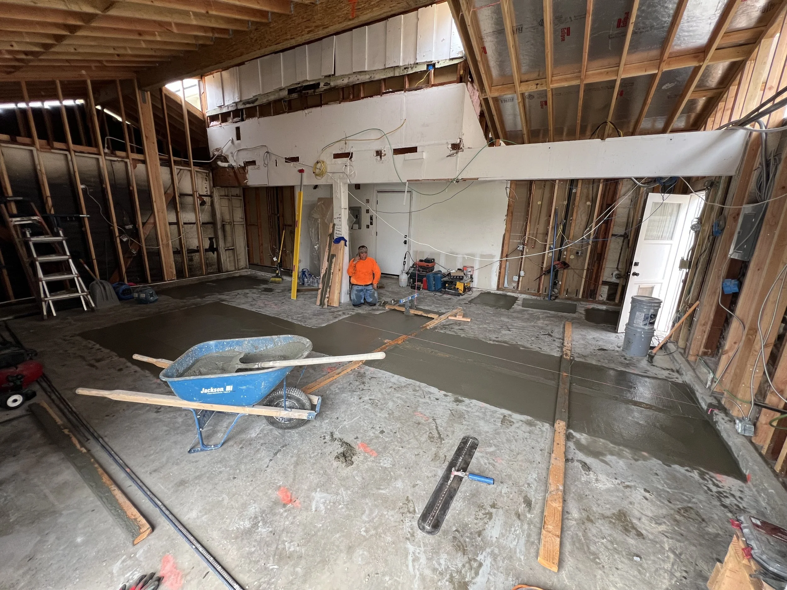 Interior of a house under renovation with concrete being poured on the floor, construction tools scattered, and a worker kneeling in an orange shirt.