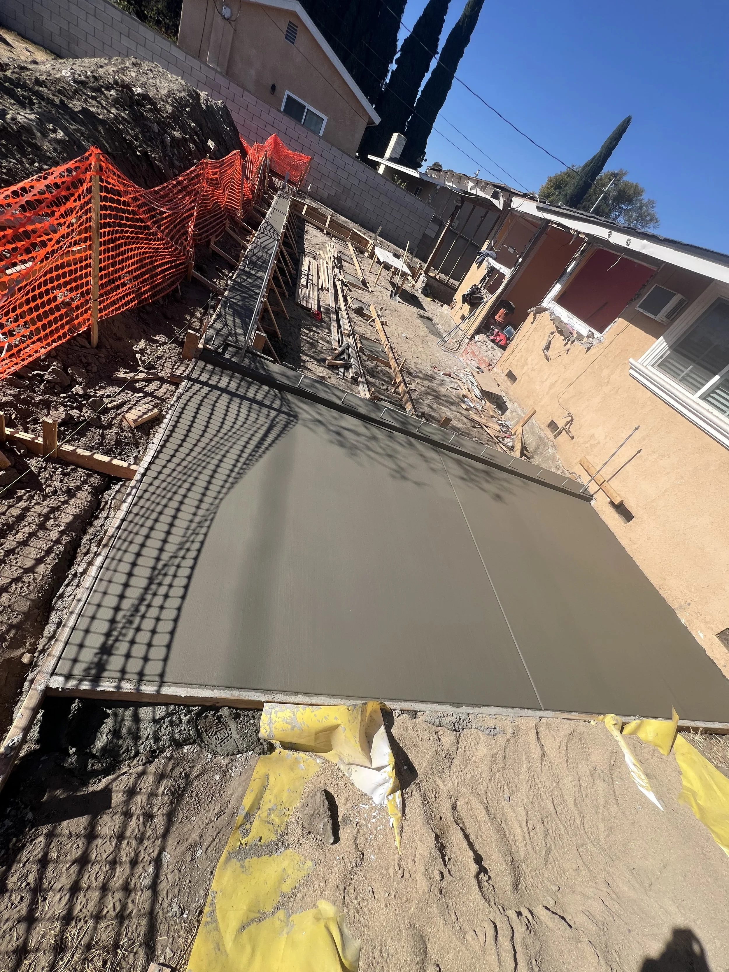 Construction site with freshly poured concrete on a pathway, fenced off with orange safety netting, in a backyard with a house and trees in the background.