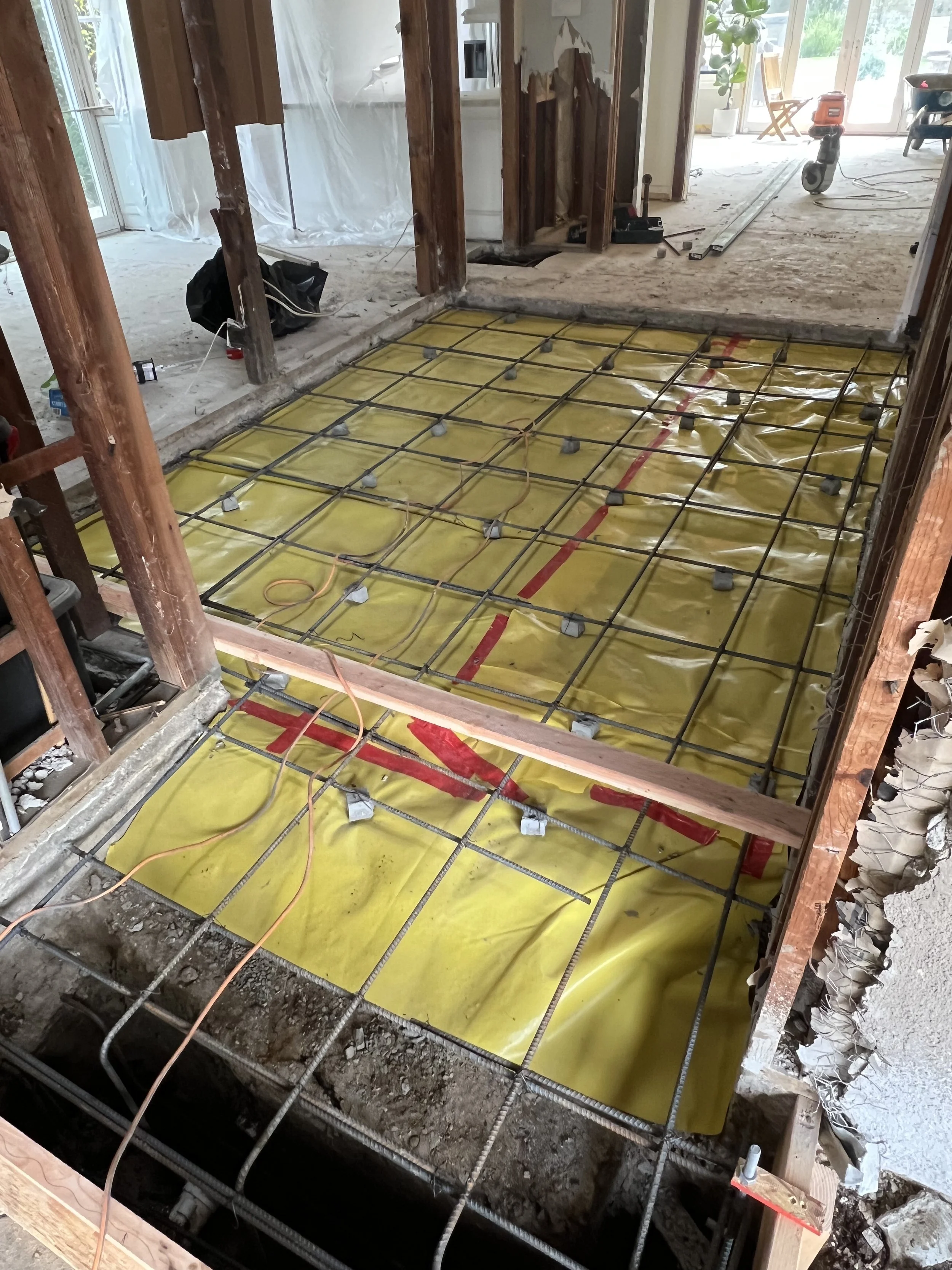 Construction site showing a room with a rebar framework for a concrete floor, yellow vapor barrier, and tools in the background.