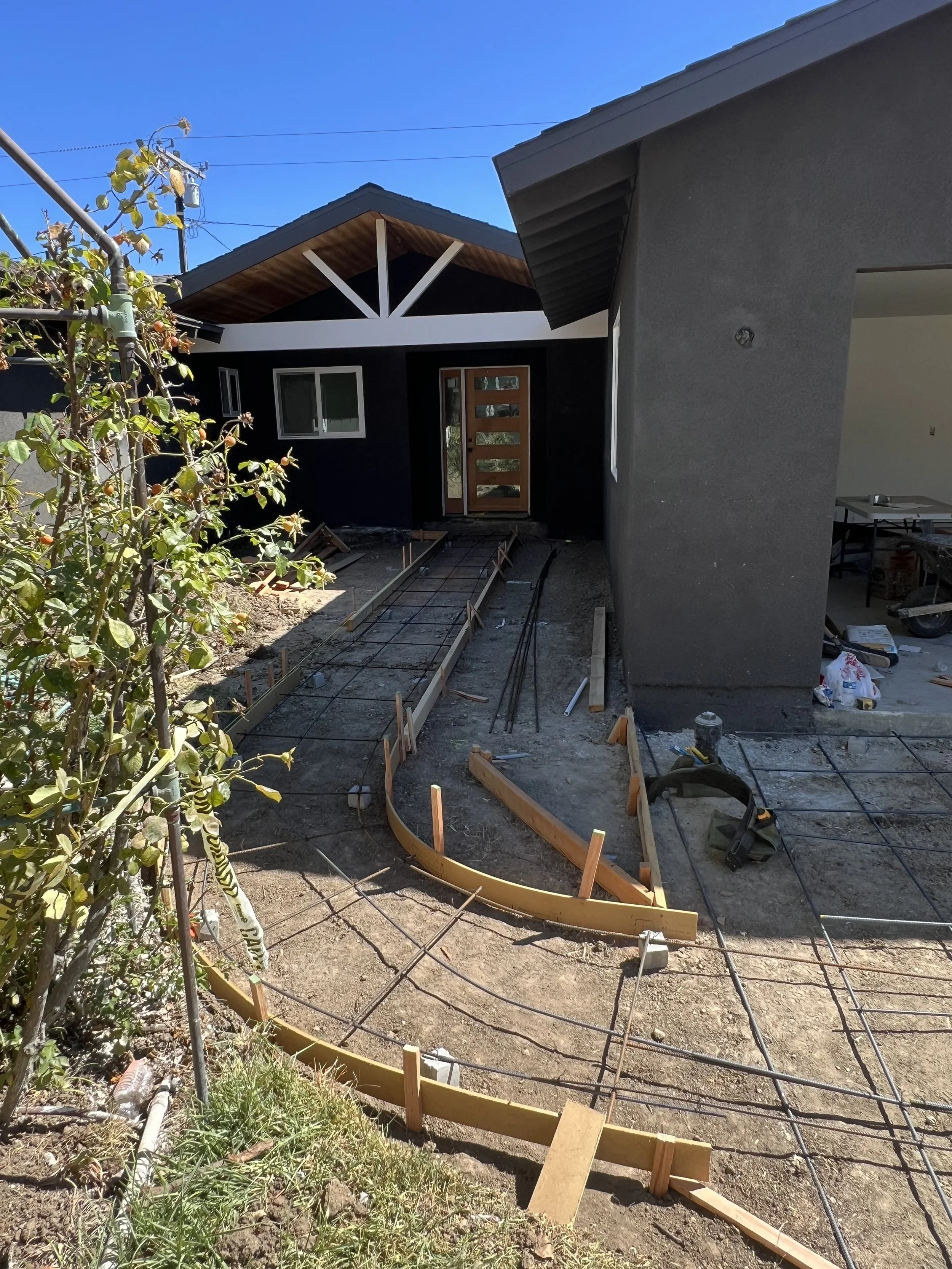 Construction site in front of a house with a new curved wooden formwork and metal rebar for a concrete sidewalk or patio. The house has a modern design with dark exterior walls and a wooden front door. There are construction tools and materials scatt