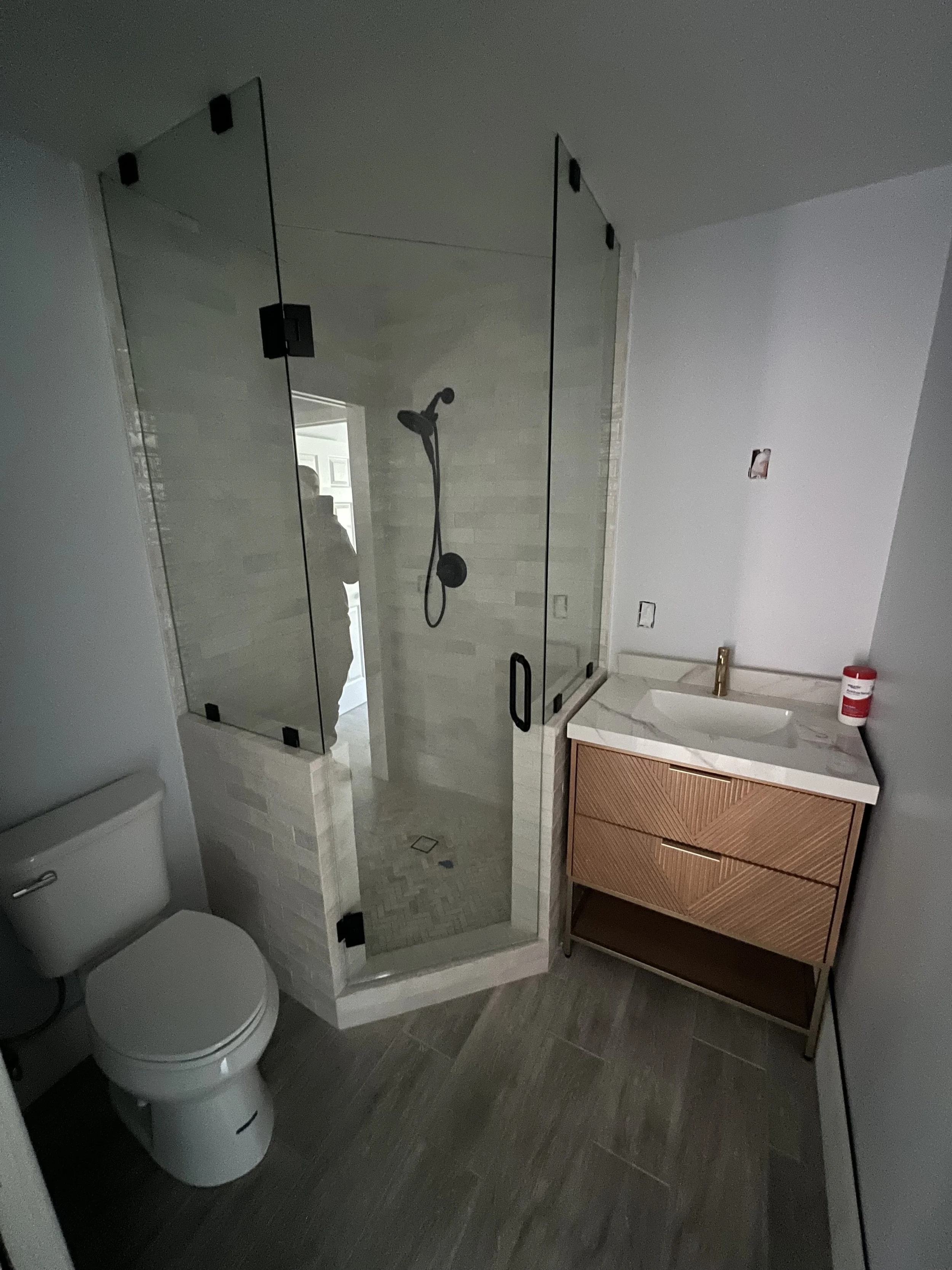 Bathroom with a glass-enclosed walk-in shower, a toilet, and a wooden vanity with a sink, with a white wall and grey tiled floor.