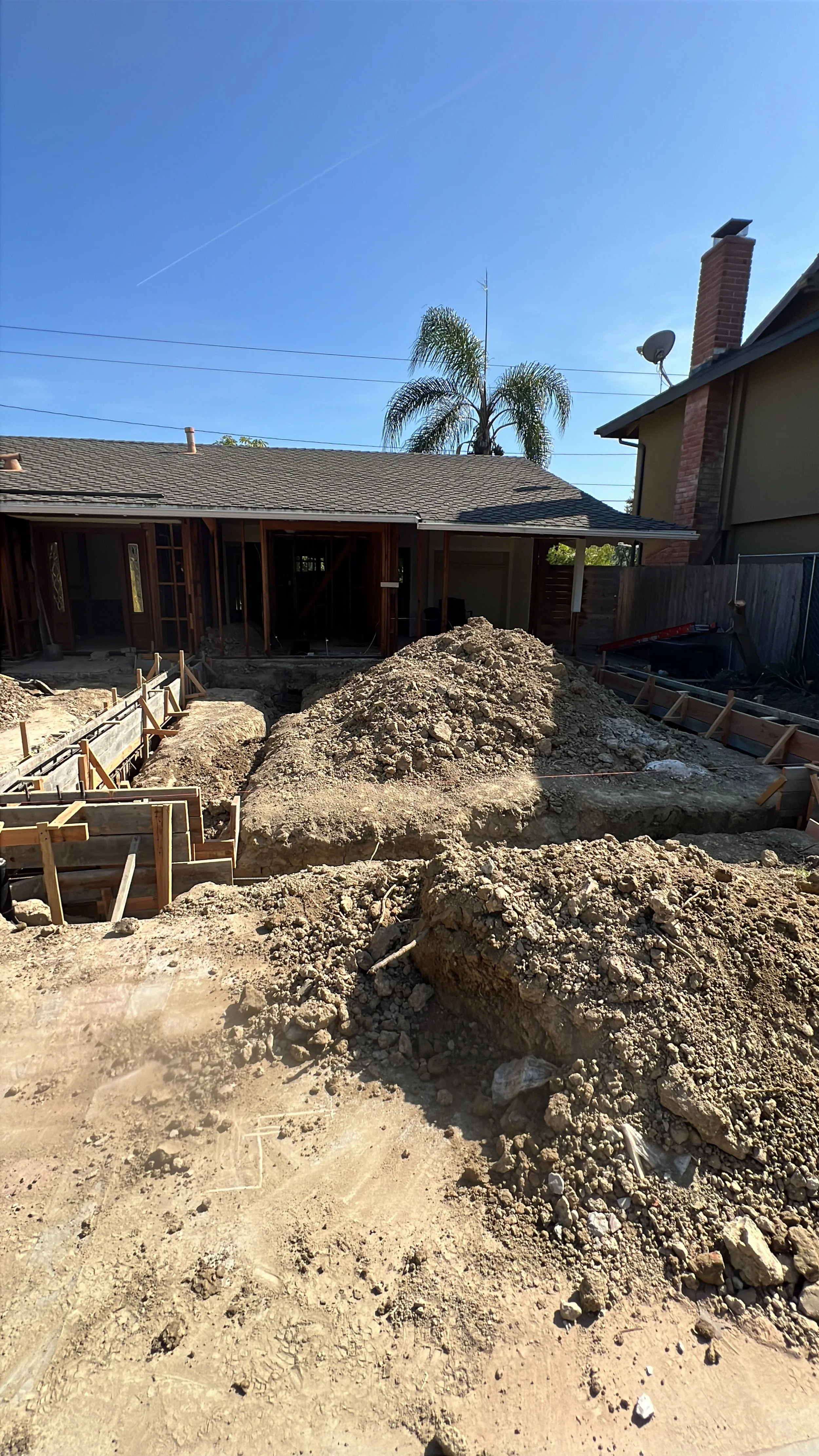 Construction site with dirt piles, trench, and wooden framing for a building project in a residential backyard.