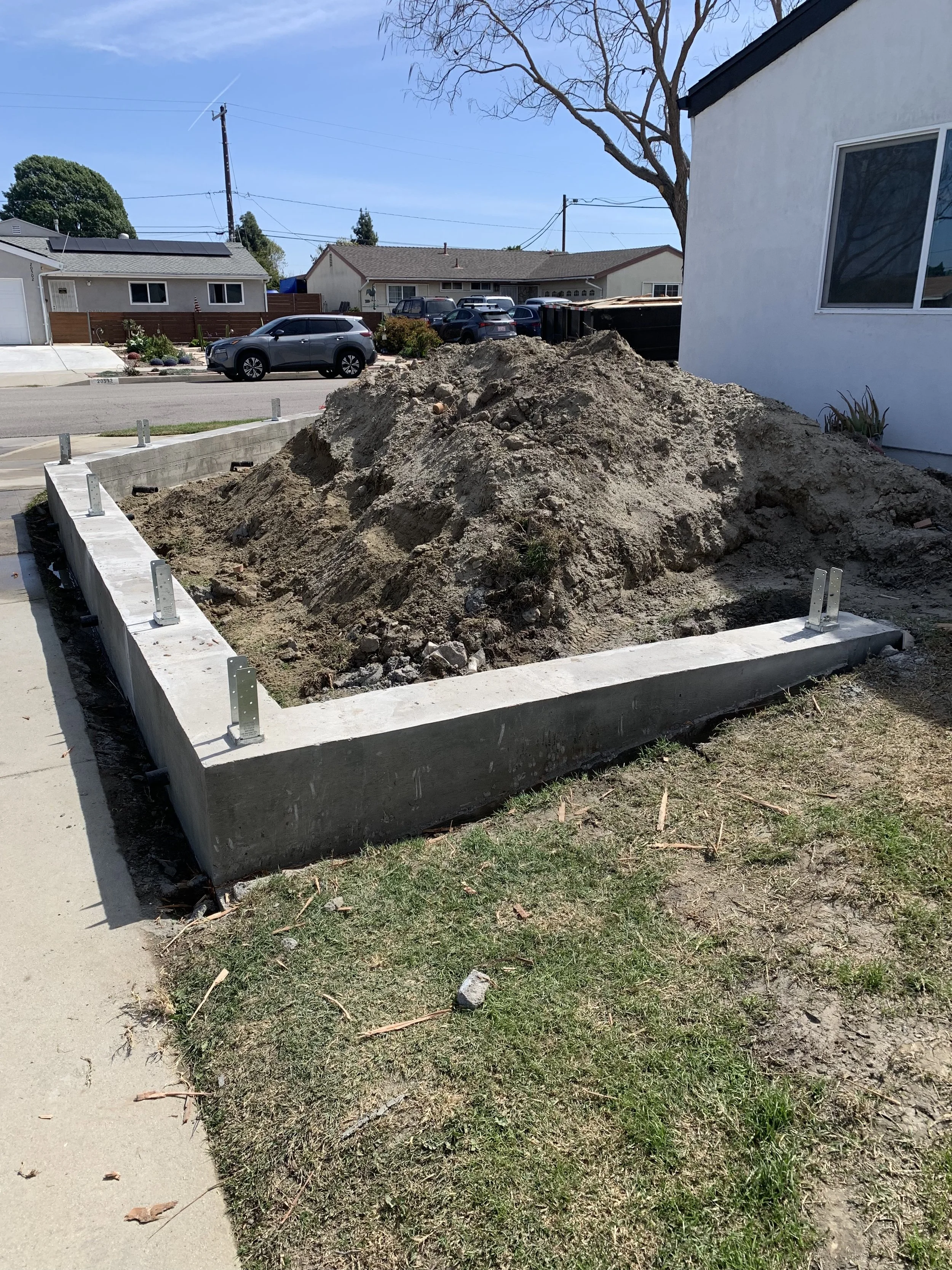 Construction site in a residential neighborhood with a concrete foundation and a pile of dirt. Houses and parked cars are visible in the background.