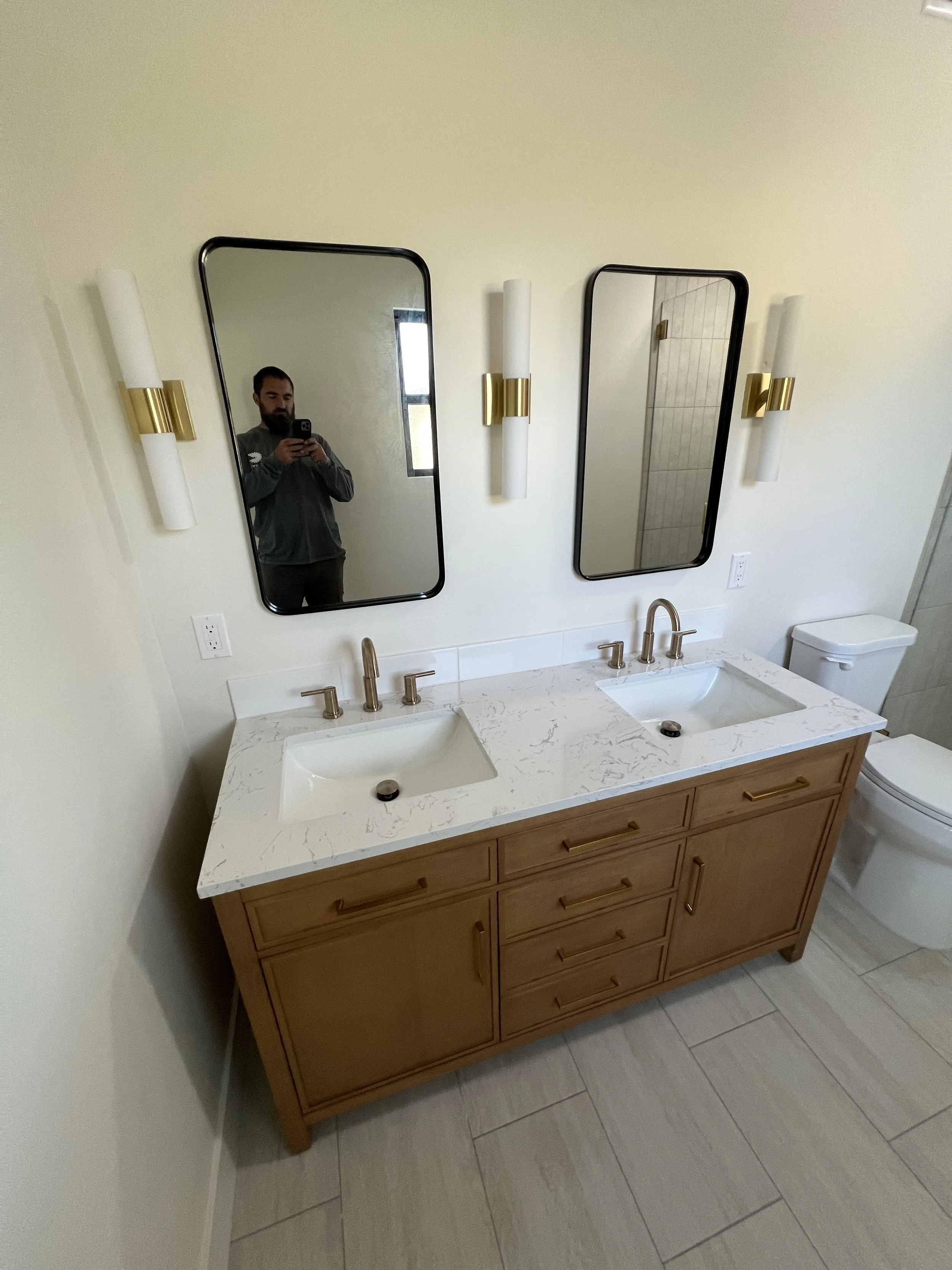 A modern bathroom with a double sink vanity, two mirrors, and wall-mounted light fixtures, with a person taking a photo with a smartphone seen in the mirror.