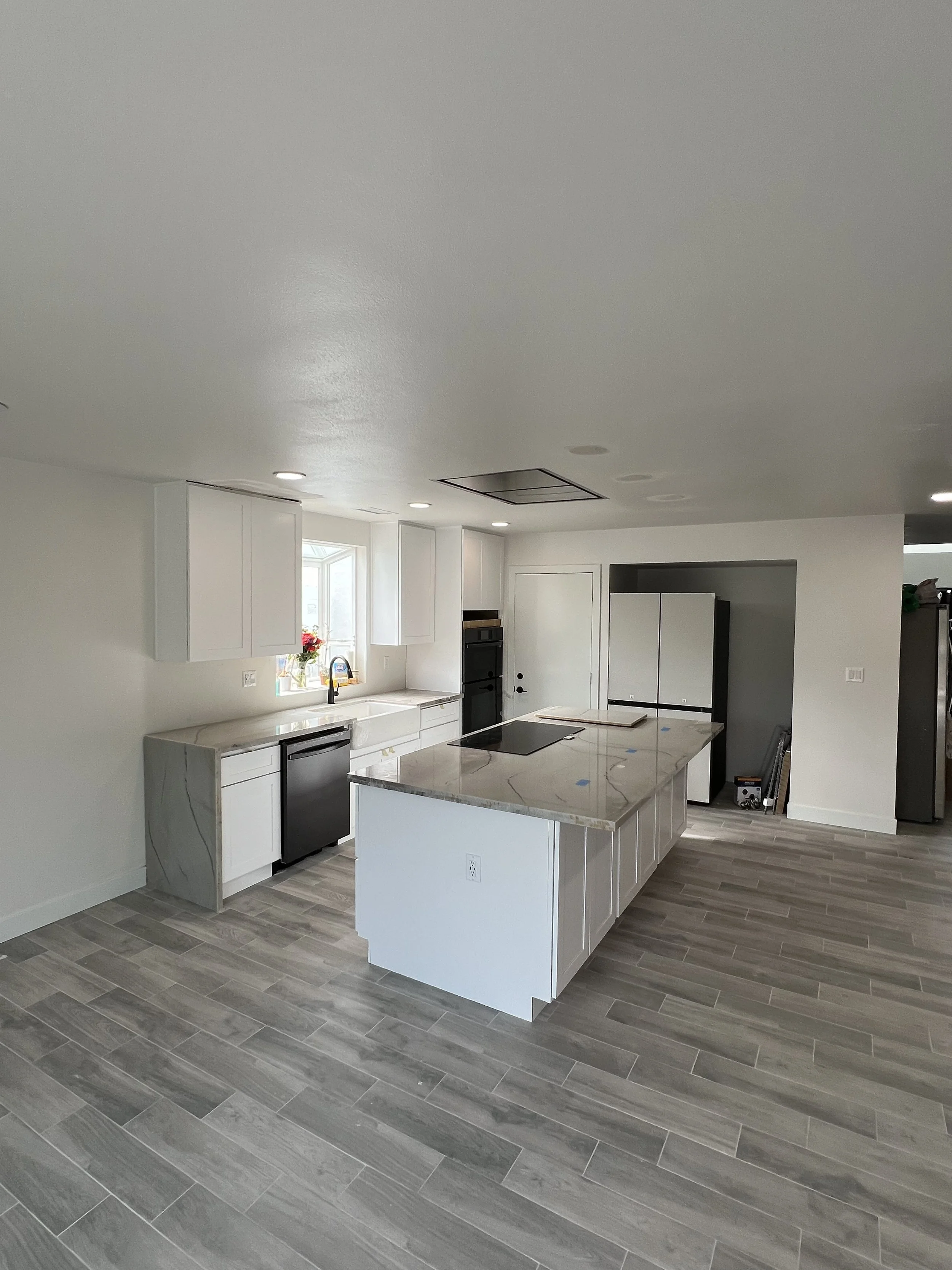 Modern kitchen with white cabinets, a large island with marble countertop, black appliances, wooden flooring, and a window with flowers on the windowsill.