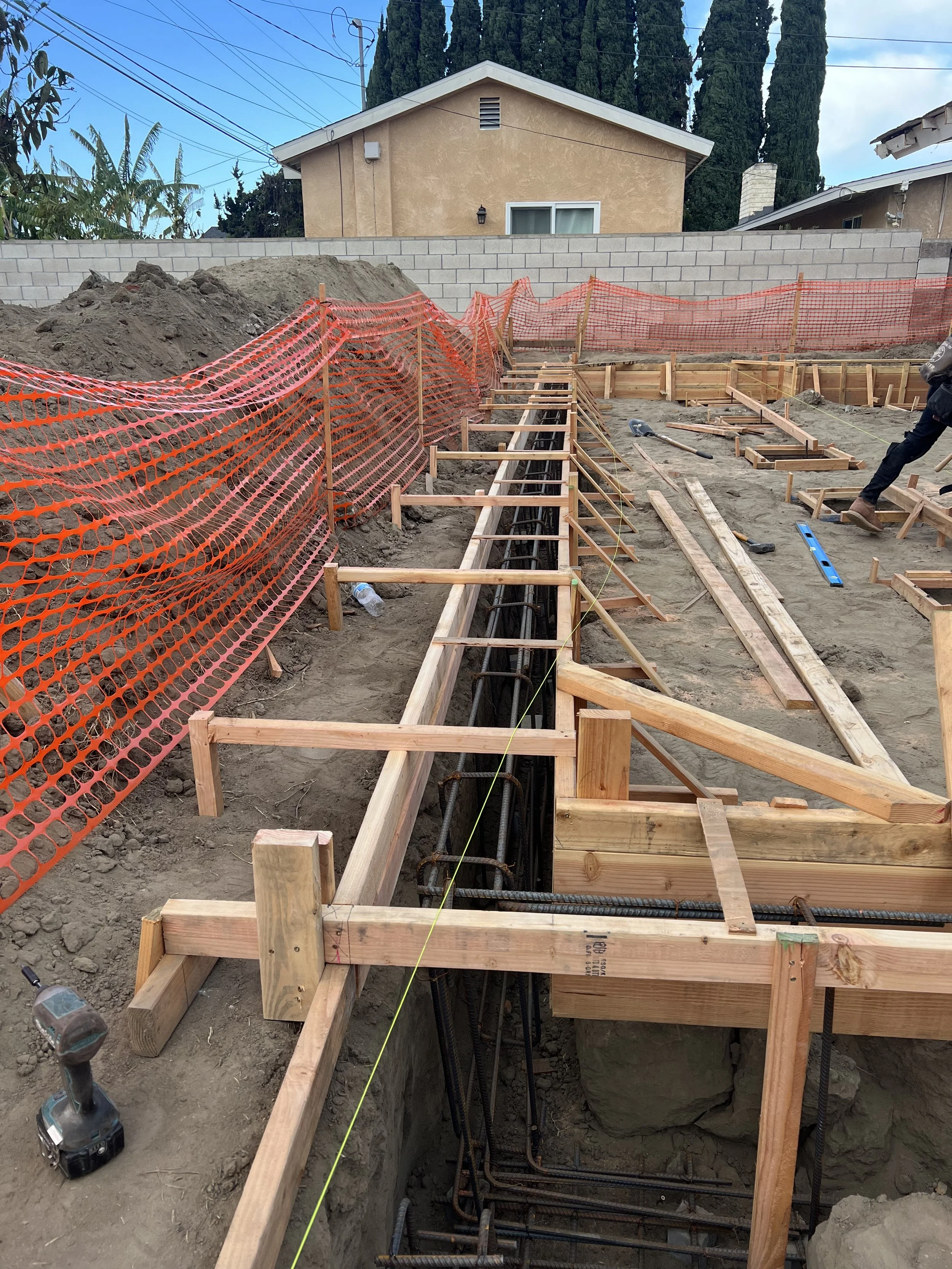 Construction site with wooden framing and rebar, orange safety fencing, and partially dug trenches for foundation work.