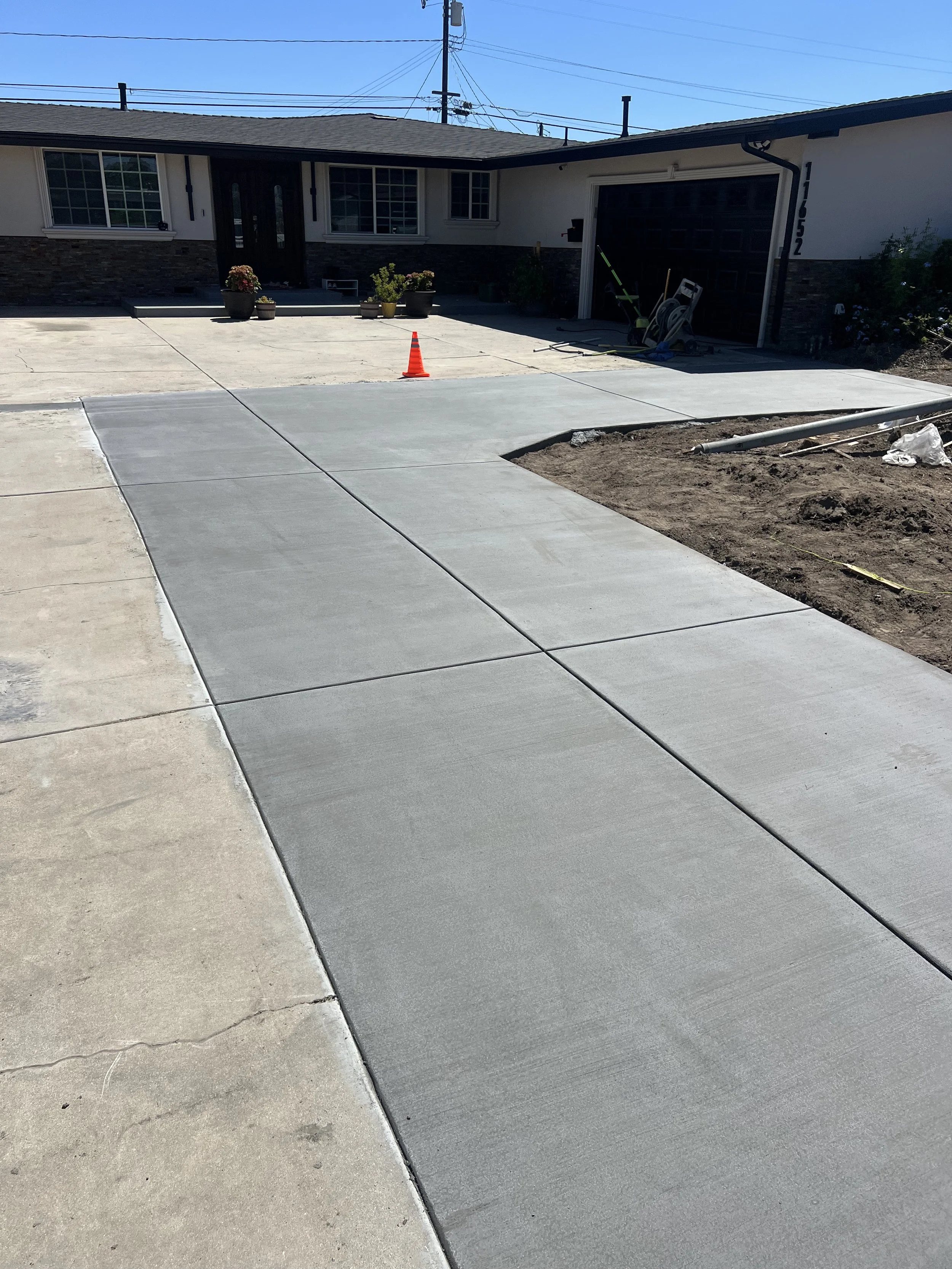 Newly poured concrete sidewalk in front of a house with two potted plants near the entrance, and construction materials and equipment nearby.