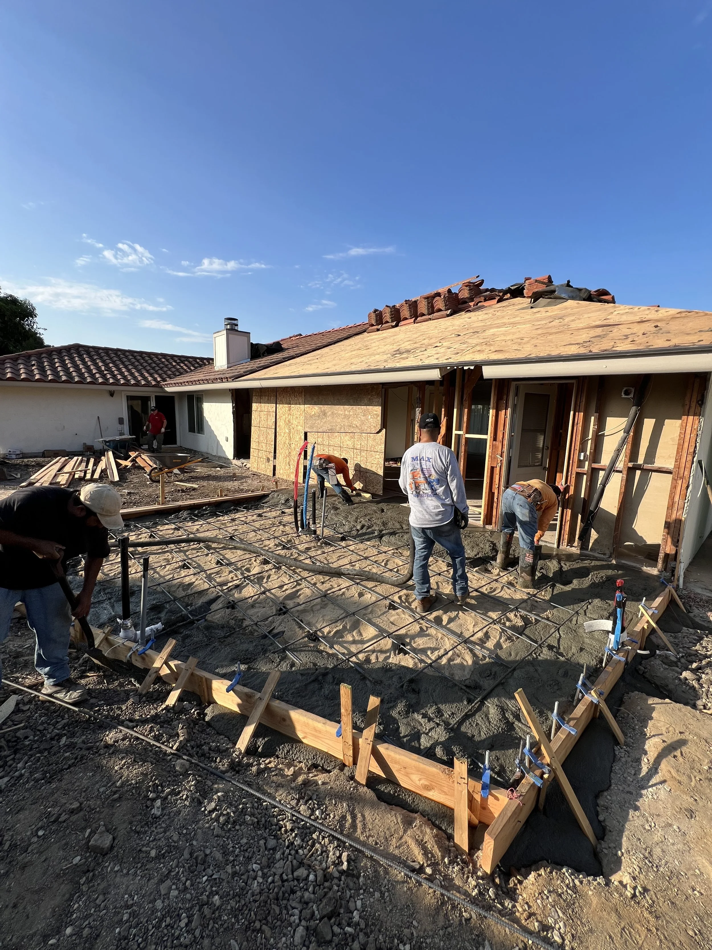 Construction workers pouring concrete on a foundation in front of a house under renovation with a clear blue sky overhead.