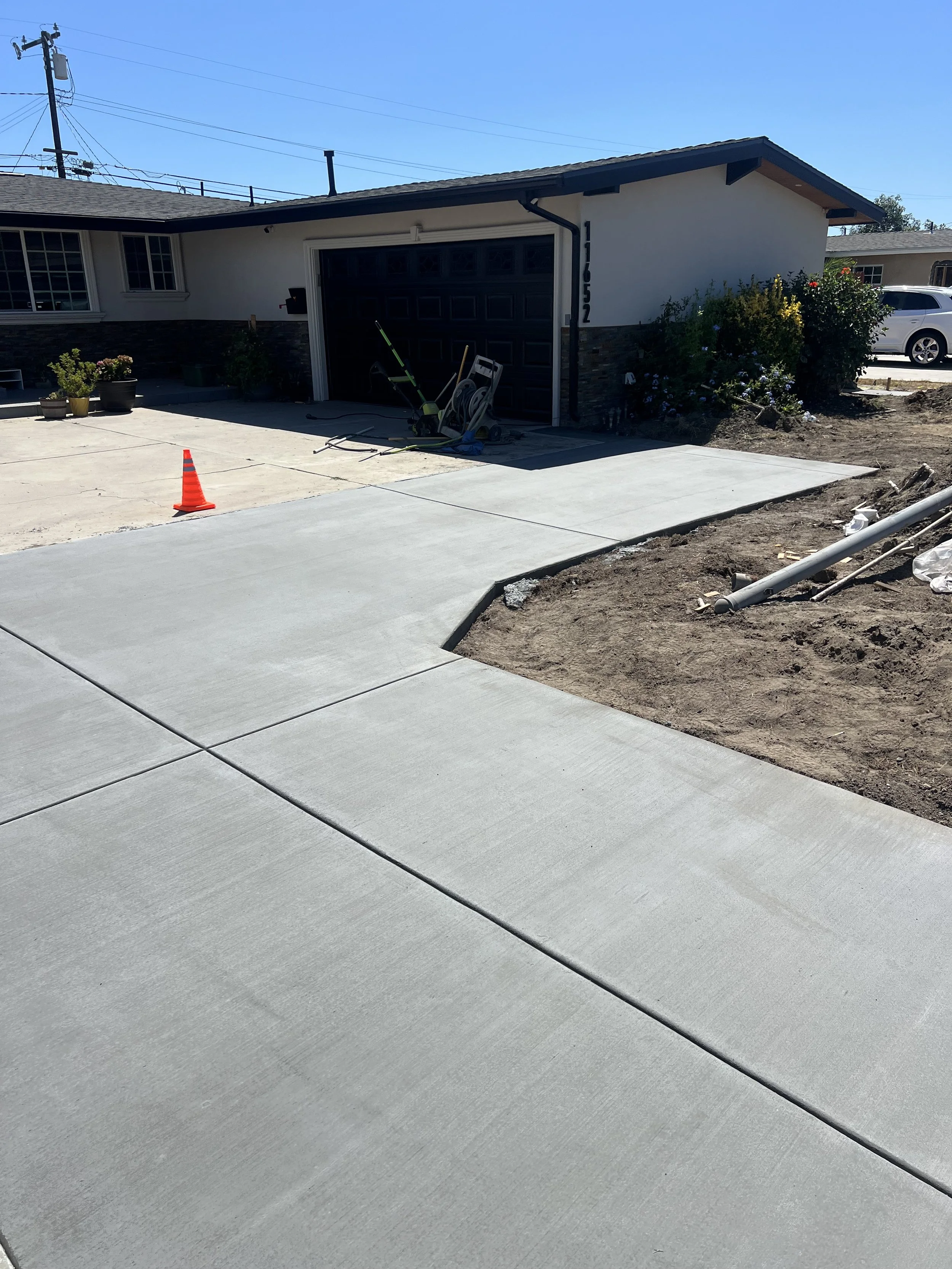Newly poured concrete driveway leading to a garage of a house under construction or renovation, with construction tools and orange safety cone nearby, and landscaping work ongoing.