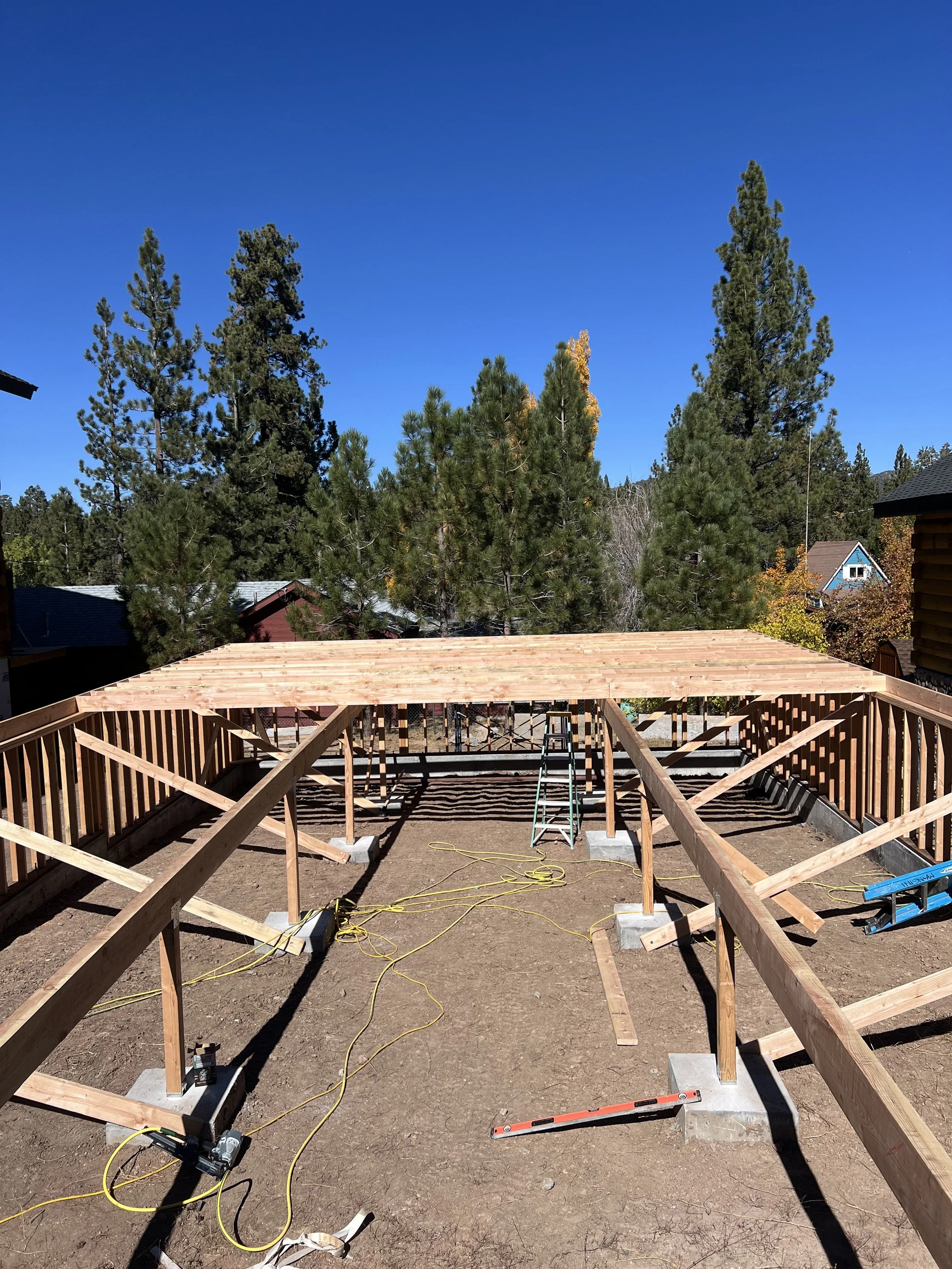 Construction site with a wooden deck frame being built, surrounded by trees under a clear blue sky.
