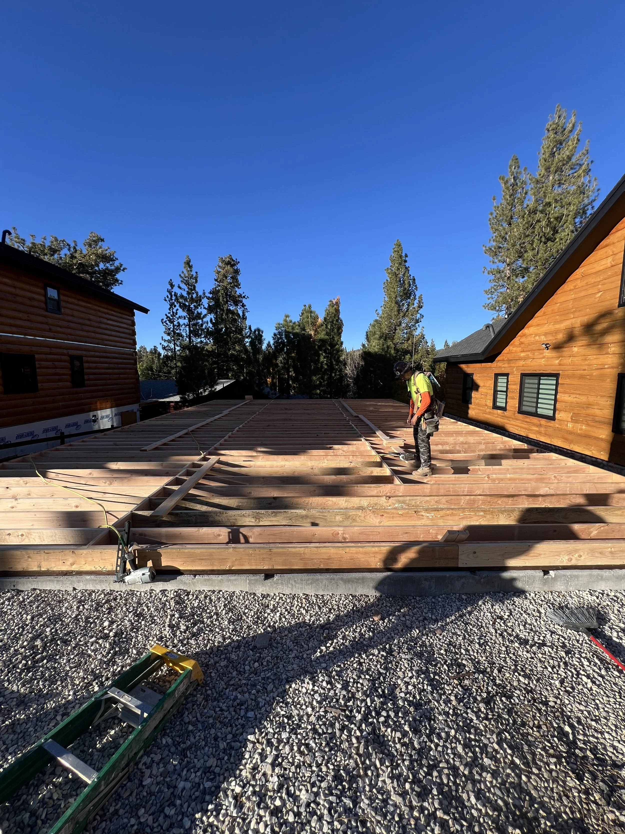 Construction worker standing on a wooden deck under construction, with a bright blue sky and pine trees in the background, on a sunny day.