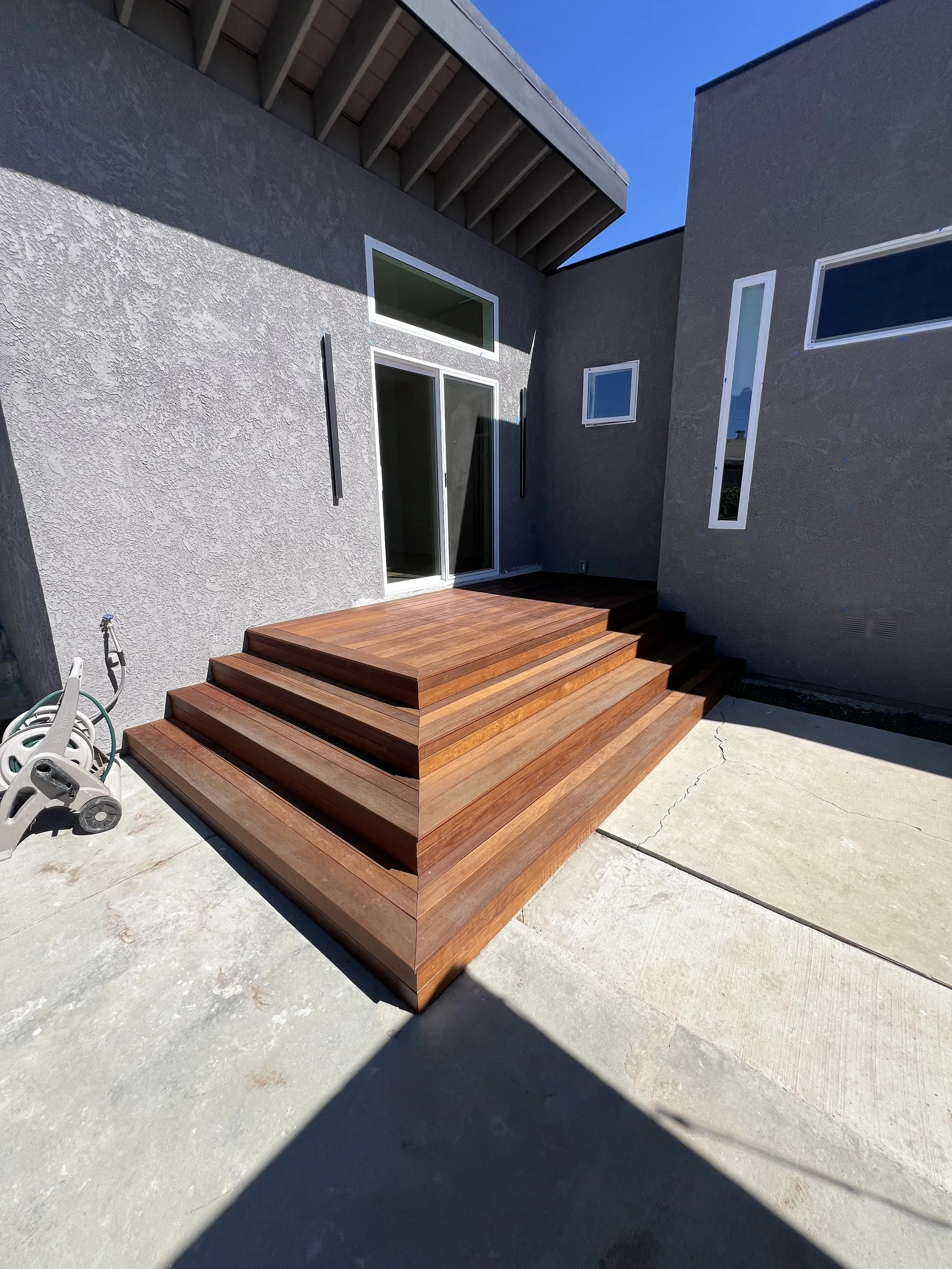 Wooden deck stairs outside a modern house with gray textured walls, multiple windows, and a concrete patio.