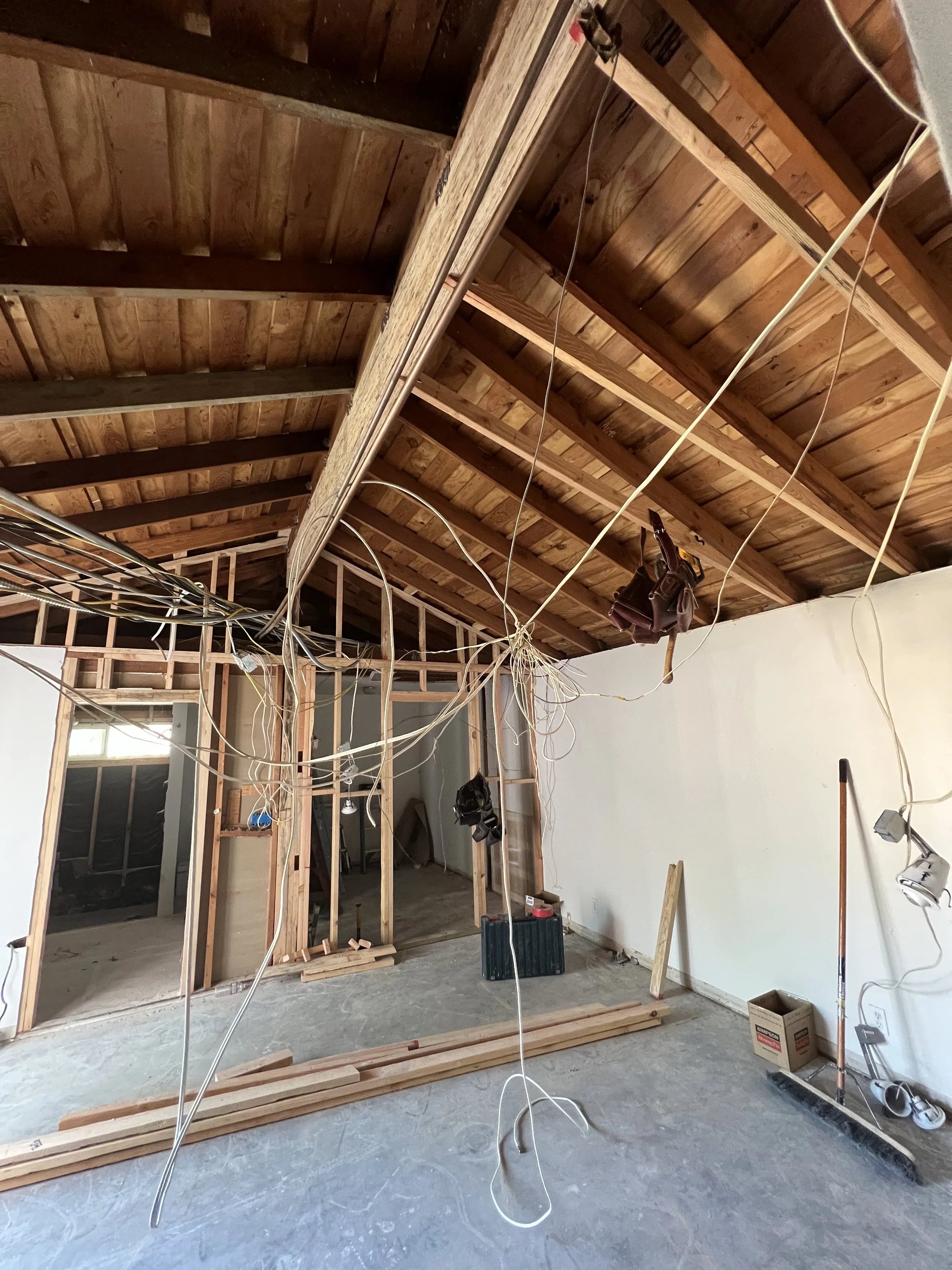Interior of a room under construction with exposed wooden framing, electrical wires hanging from the ceiling, and construction tools and materials on the floor.