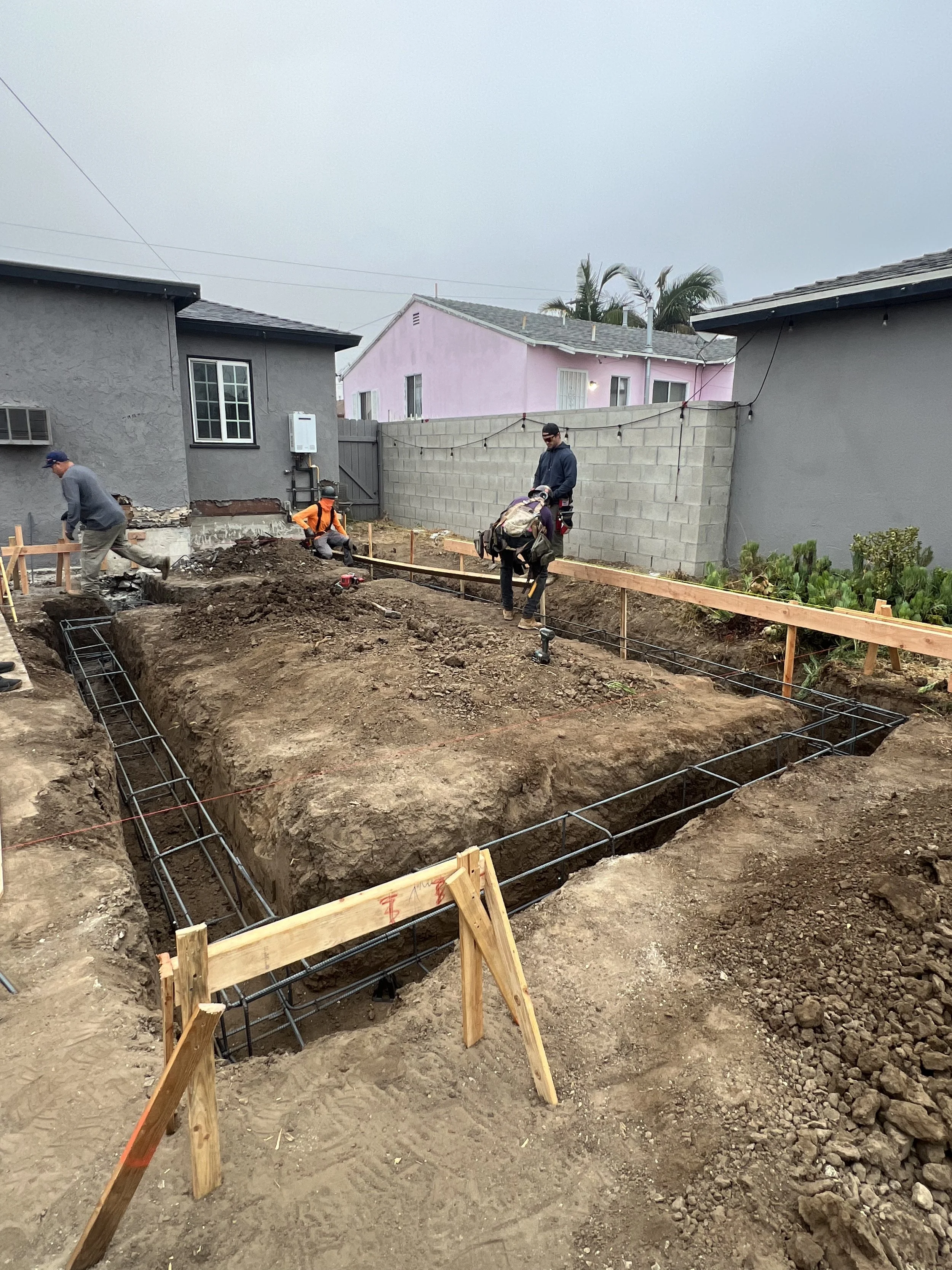 Construction workers preparing the foundation of a building, with rebar and wooden markings, in a residential backyard with neighboring houses and a gray sky.