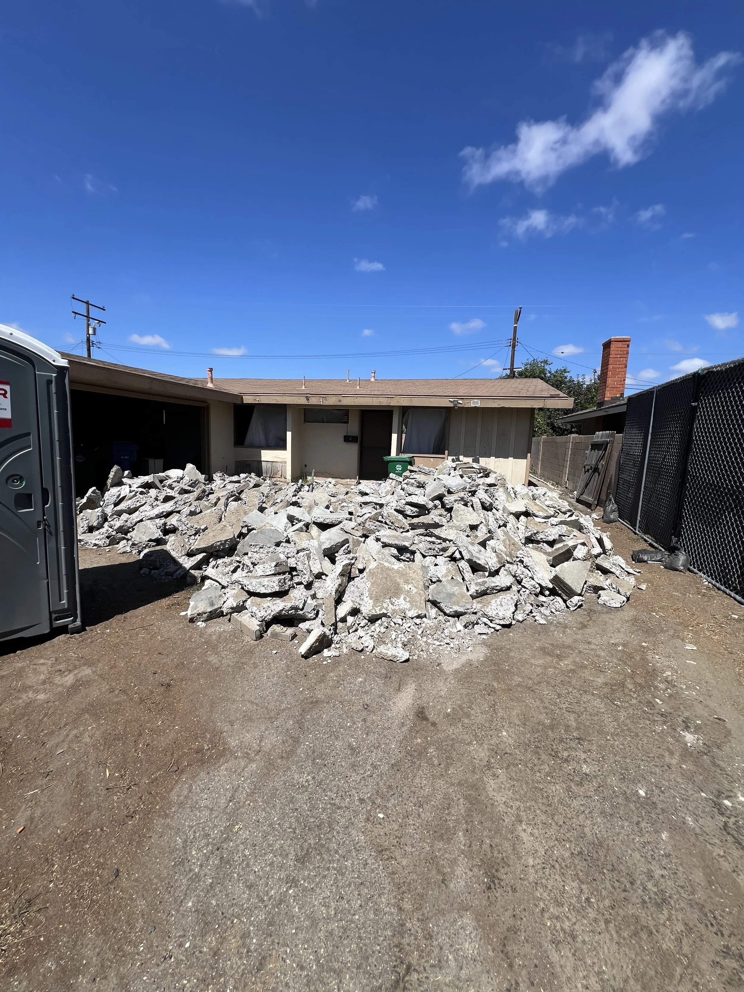 Pile of gray rocks in a backyard with a house and clear blue sky in the background.