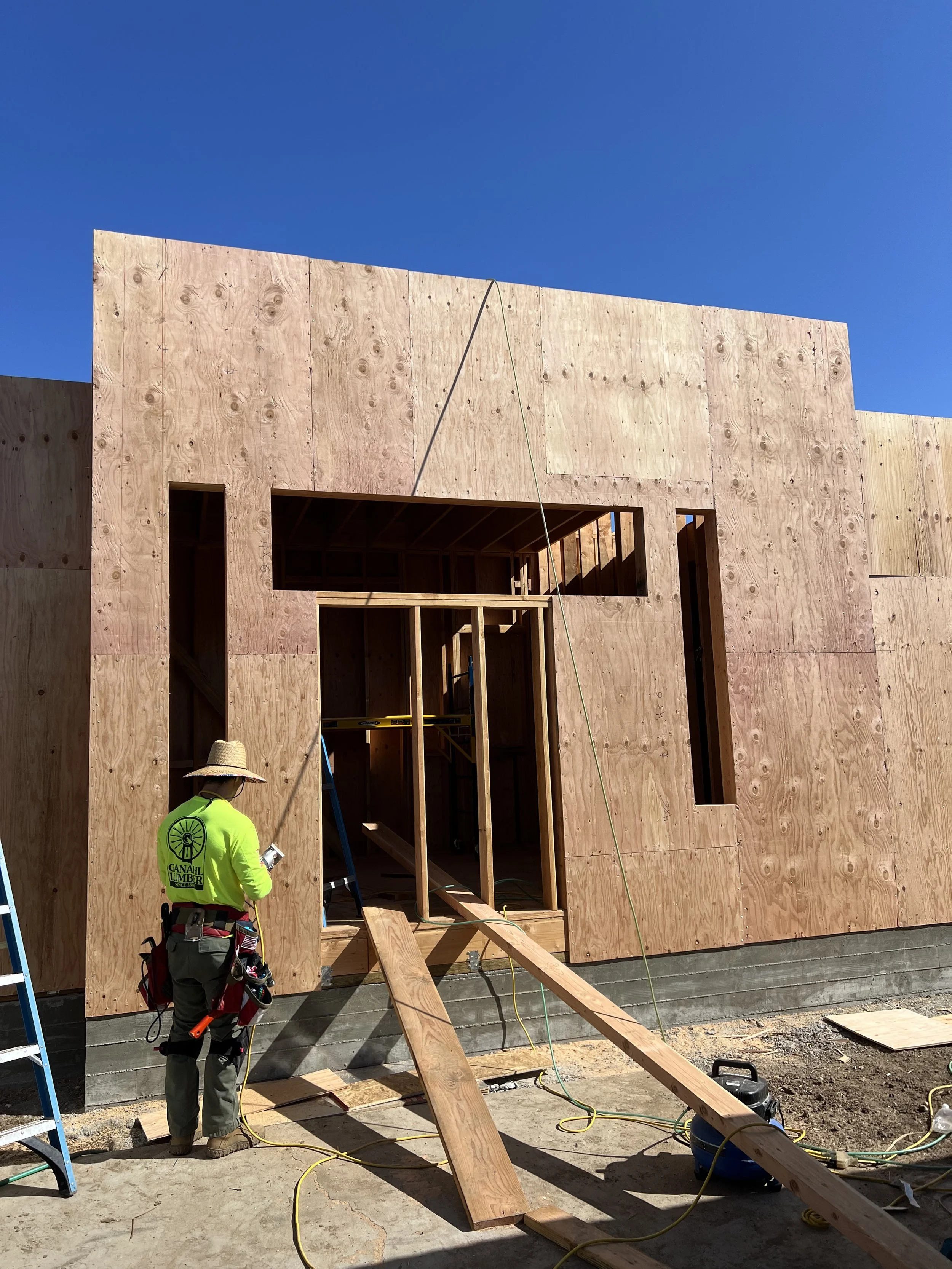 Construction worker in safety gear inspecting a partially built wooden house under a clear blue sky.