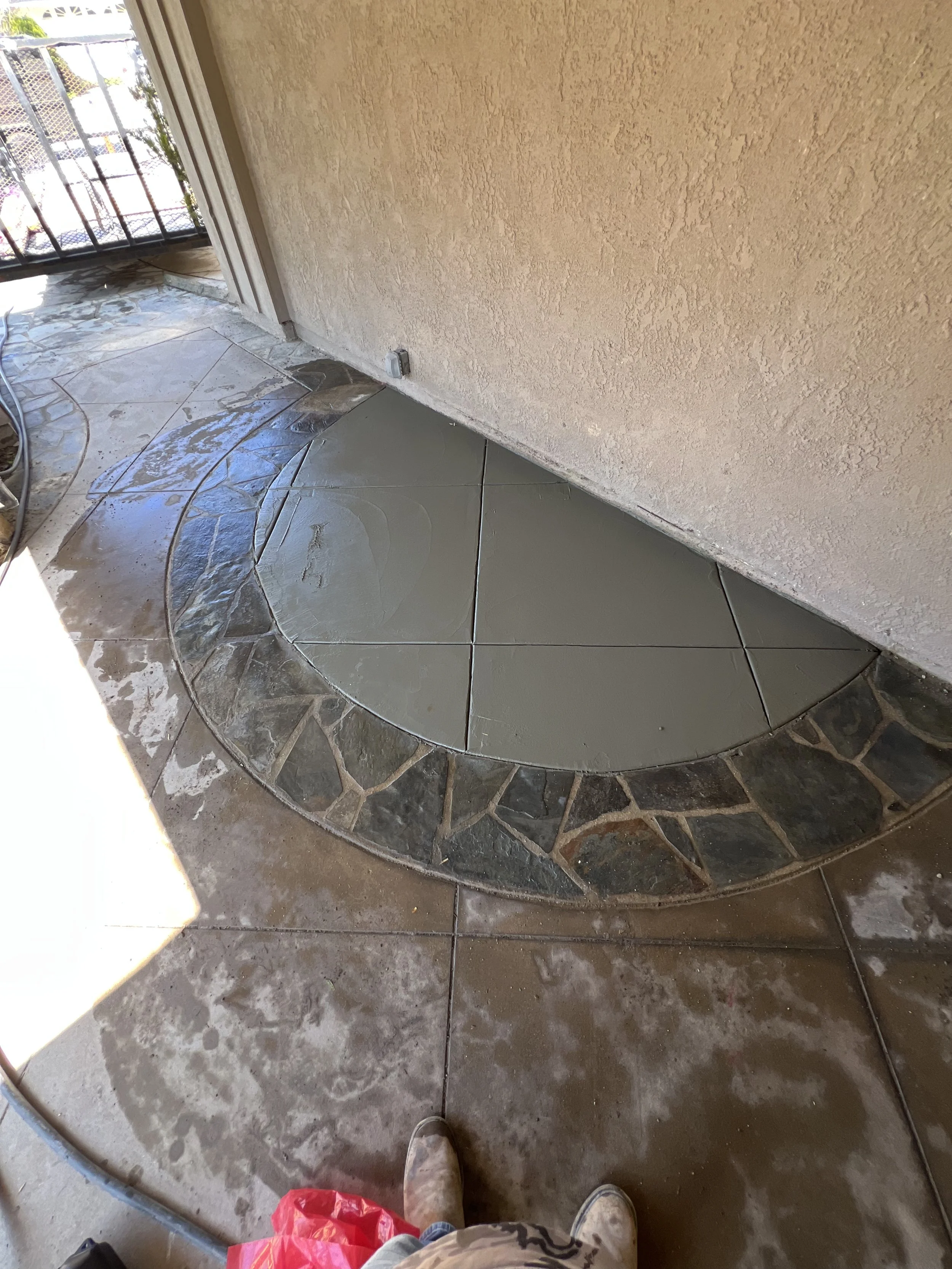 A partially completed decorative tile pathway outside a building with a beige stucco wall and a metal railing in the background. The pathway has a central section of gray tiles and a surrounding border of irregularly shaped dark stones.