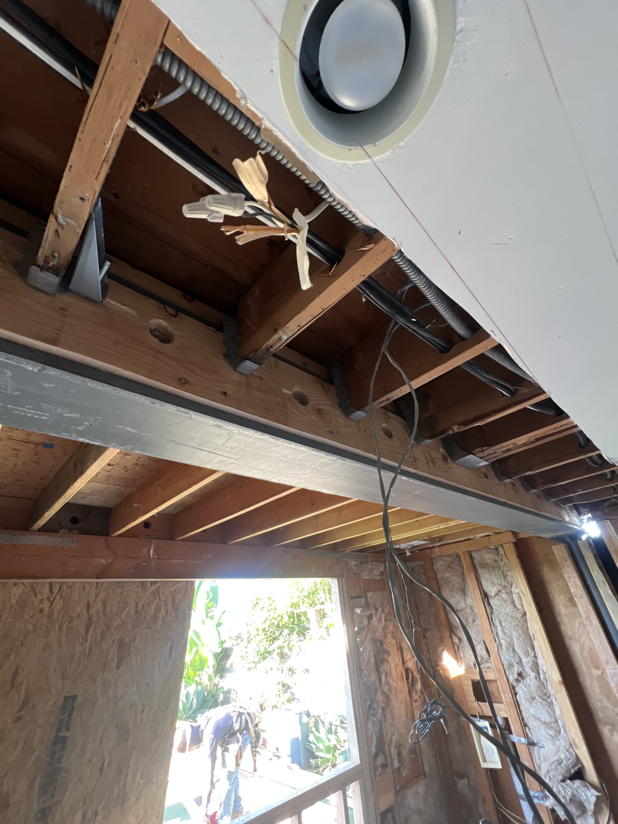 Ceiling open construction area with electrical wires, metal support beams, and roof joists, near a window with plants outside.