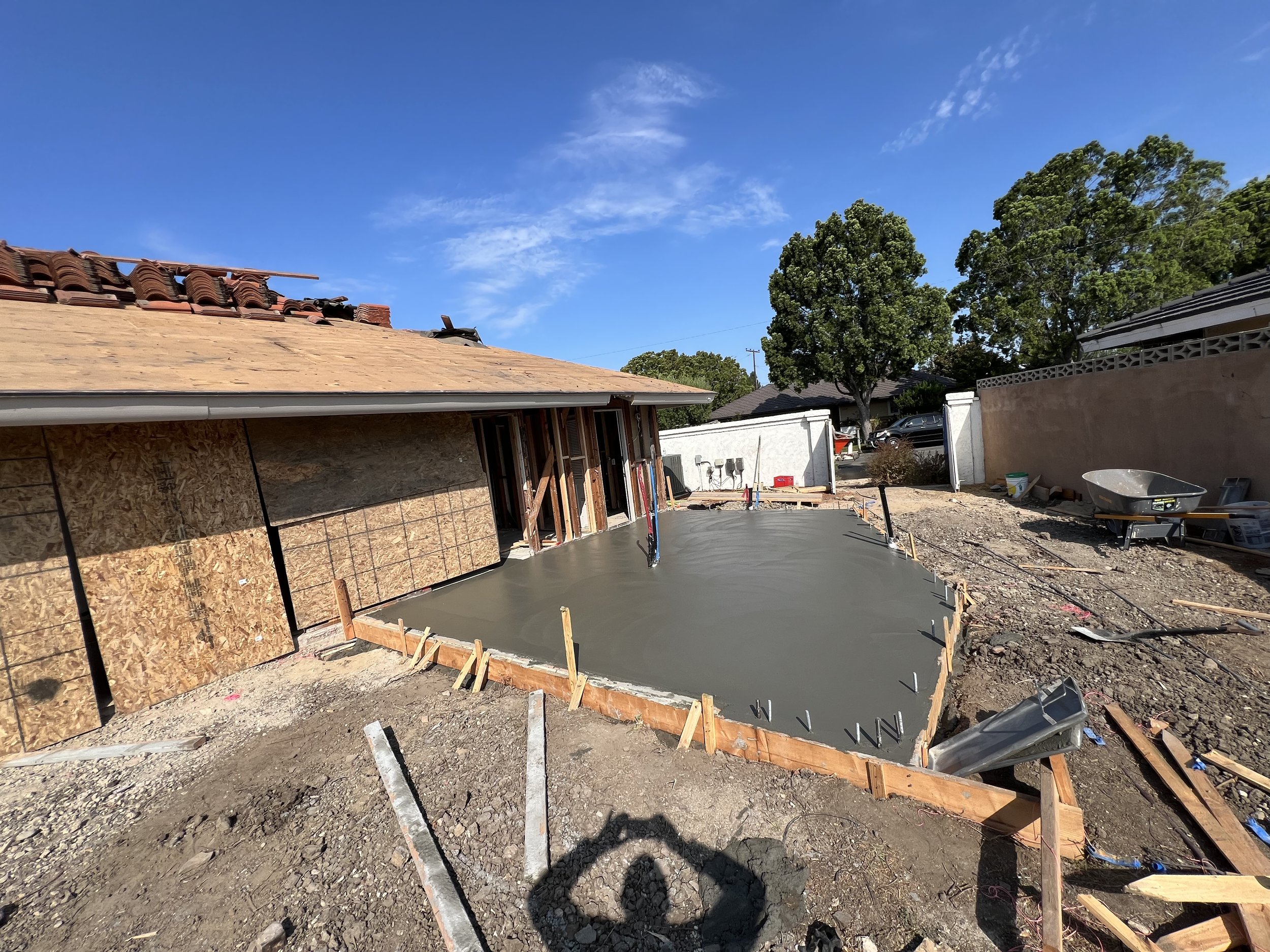 Concrete foundation being poured in a backyard construction site, with partially completed house wall and trees in the background under a clear blue sky.