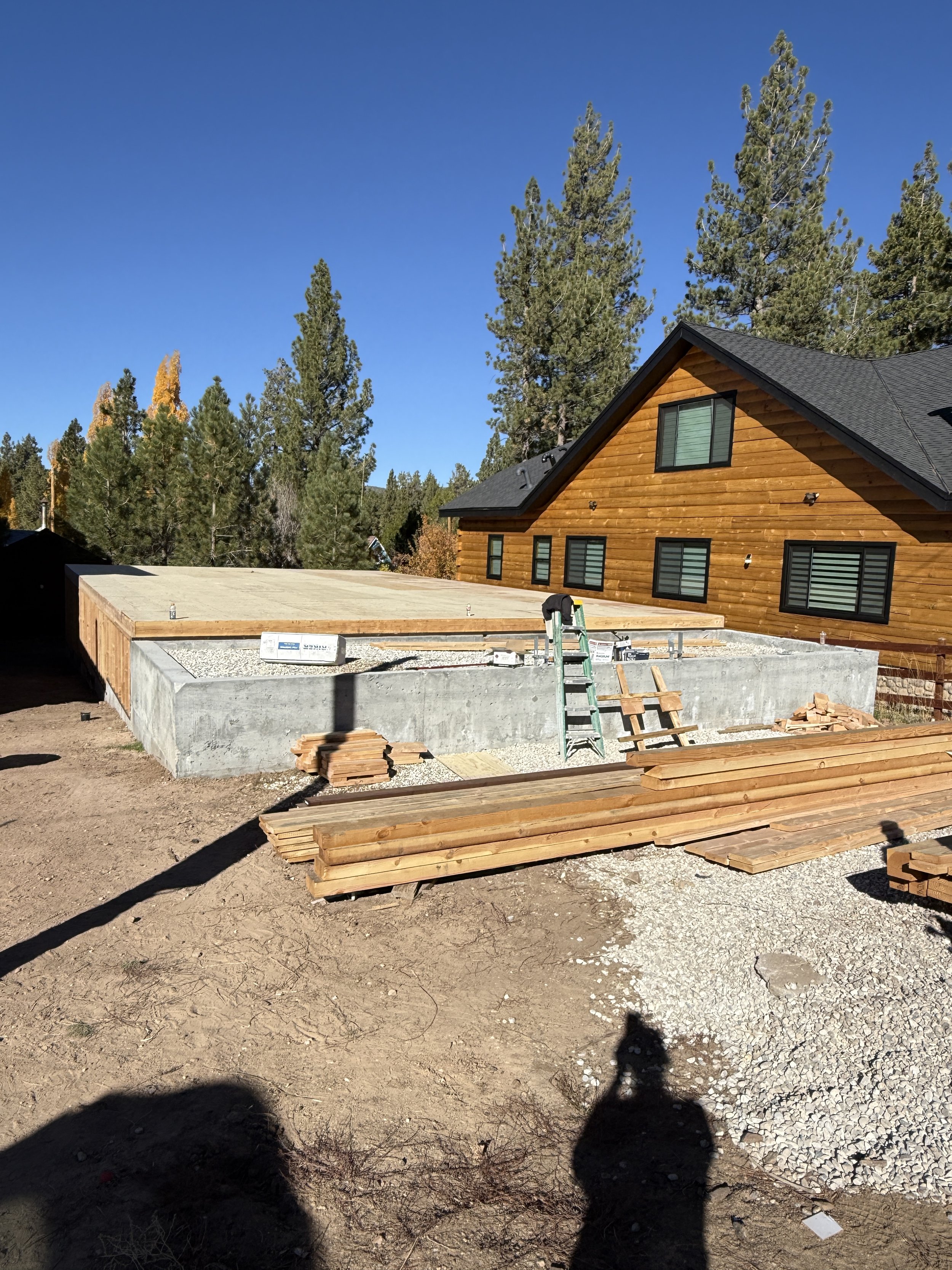 Construction site with a house foundation, wooden planks, and a ladder set up. Trees and a blue sky in the background.