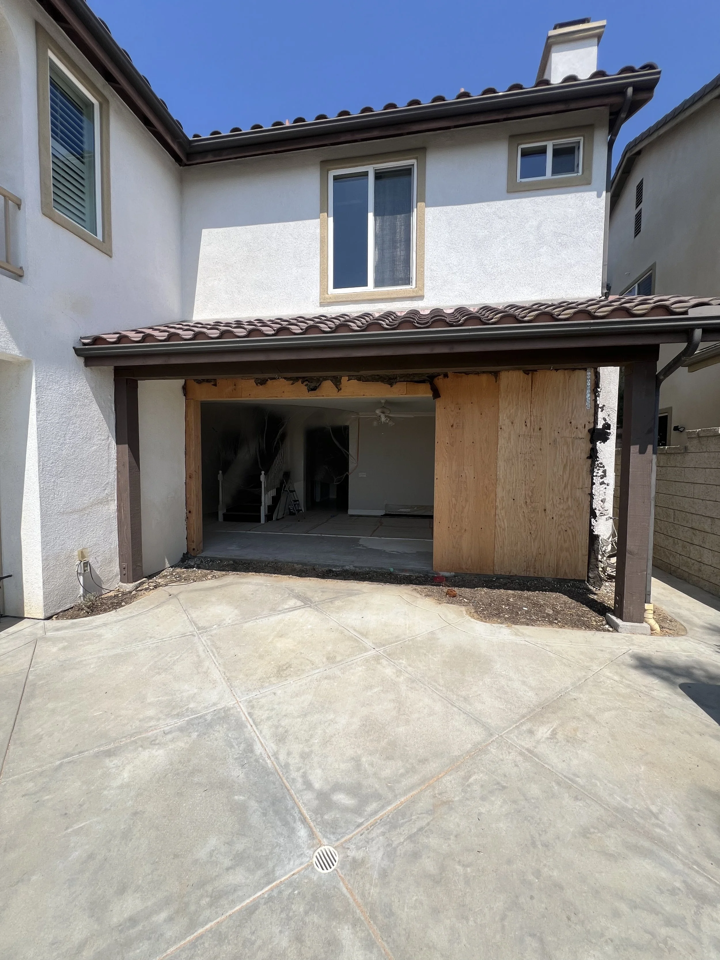 Newly constructed or renovated garage with plywood and support beams, and parts of the interior exposed. The house has a white stucco exterior with a tiled roof and nearby windows. The driveway is concrete with a drain in the center.