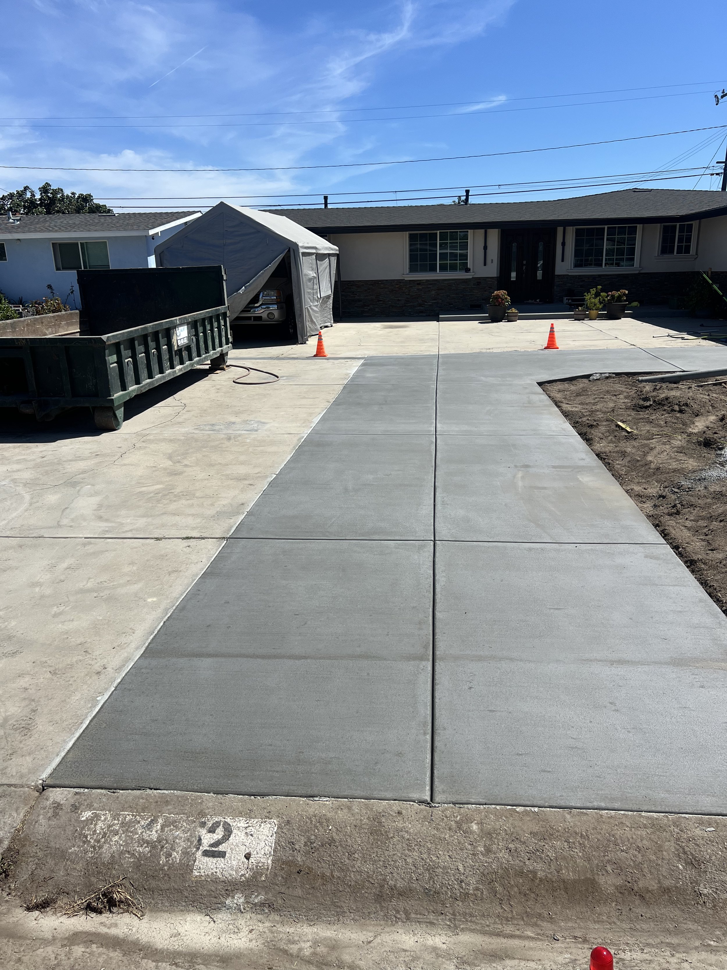 Newly poured concrete sidewalk and driveway in front of a house under construction, with orange traffic cones, a tent for construction equipment, and landscaping in progress.