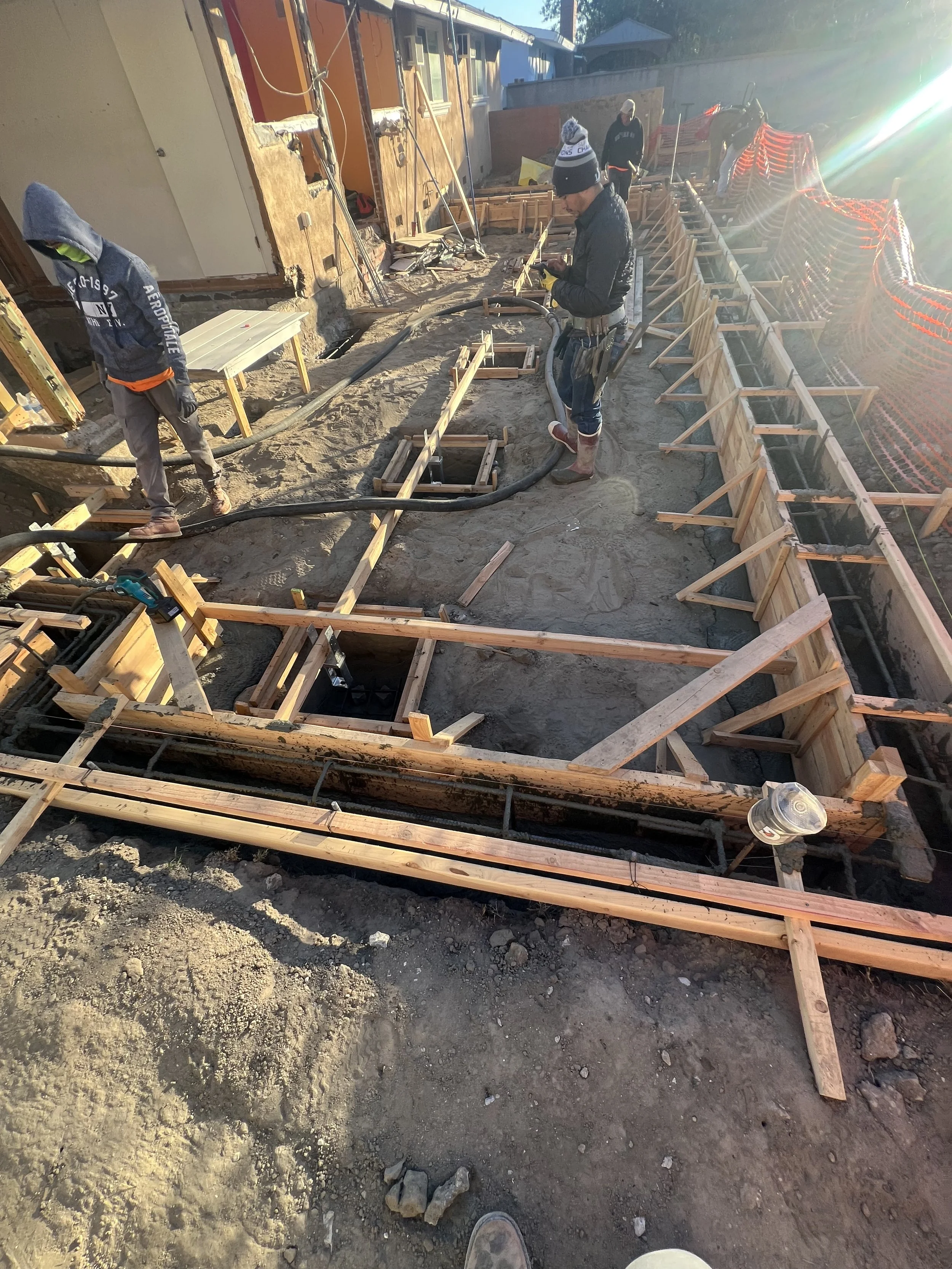 Construction workers working on a building foundation, with wooden formwork and concrete conduits, on a dirt site with sunlight.