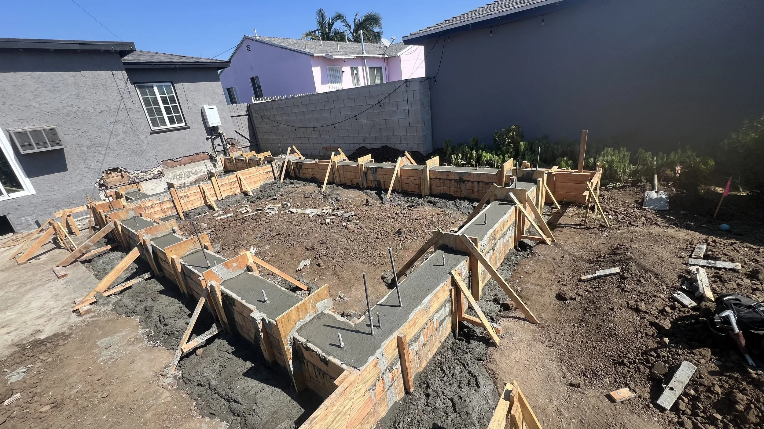 Construction site with wooden formwork and concrete for new building foundation, surrounded by dirt, in a residential backyard with neighboring houses and trees in the background.