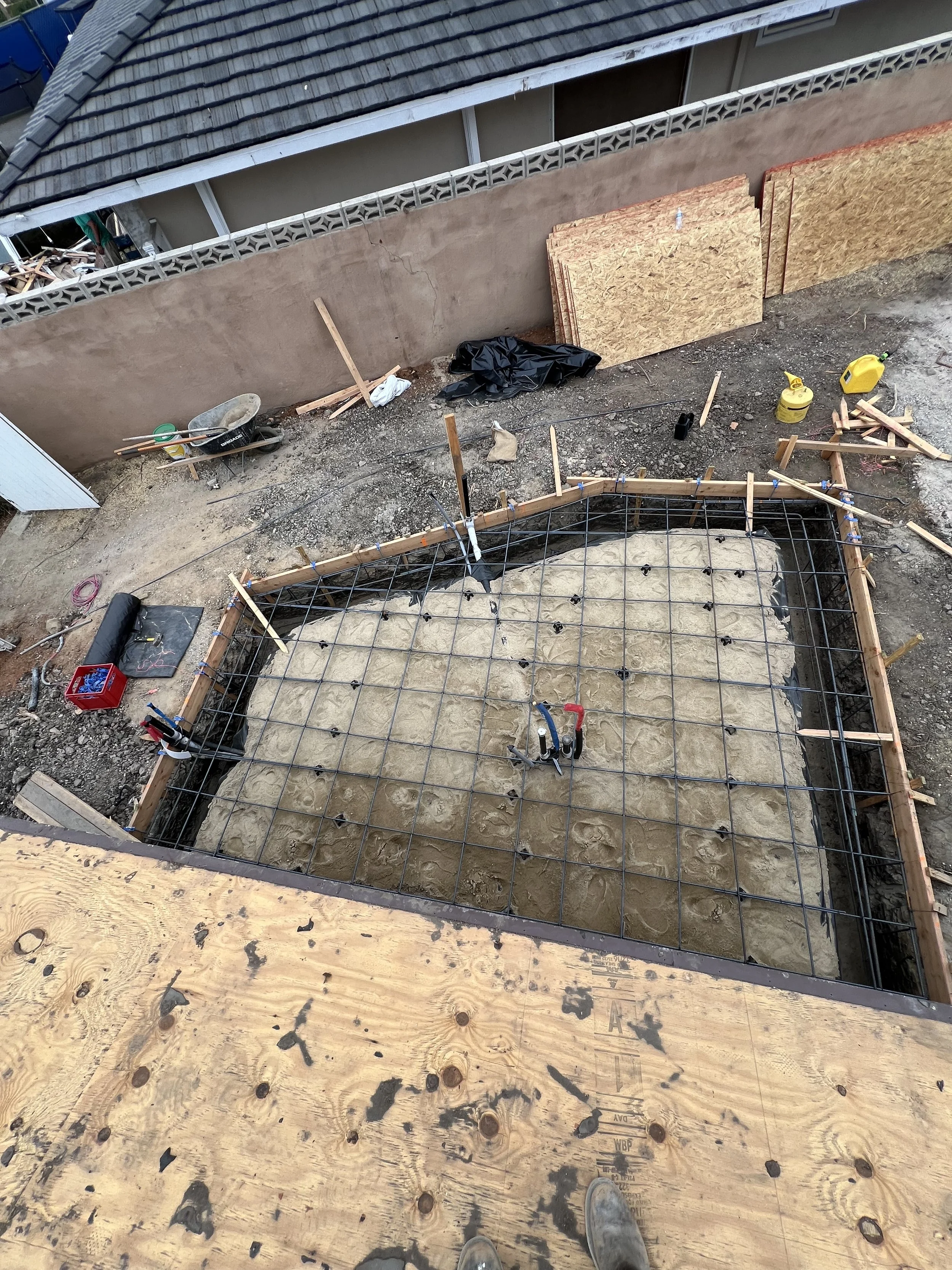 Construction site with a rectangular foundation with concrete pour and a metal rebar grid, surrounded by wooden framing, tools, and building materials. The photo is taken from a high angle.