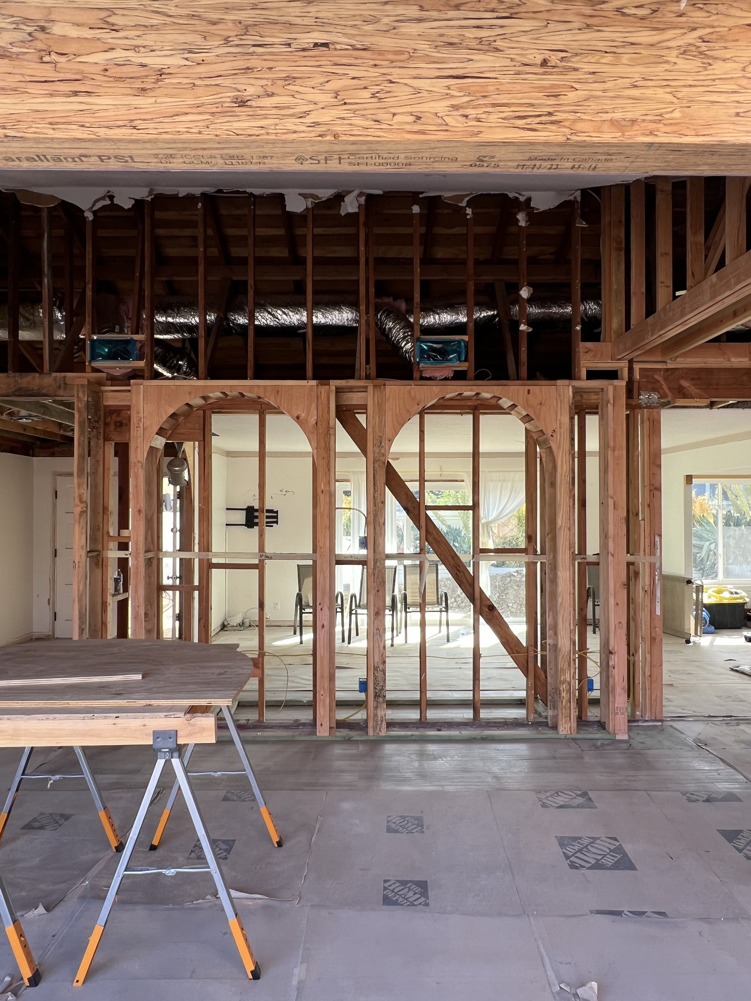 Interior view of a house under construction, showing a framed wall with arches, exposed wooden studs, a ceiling with ductwork, and some construction tools and materials on the floor.