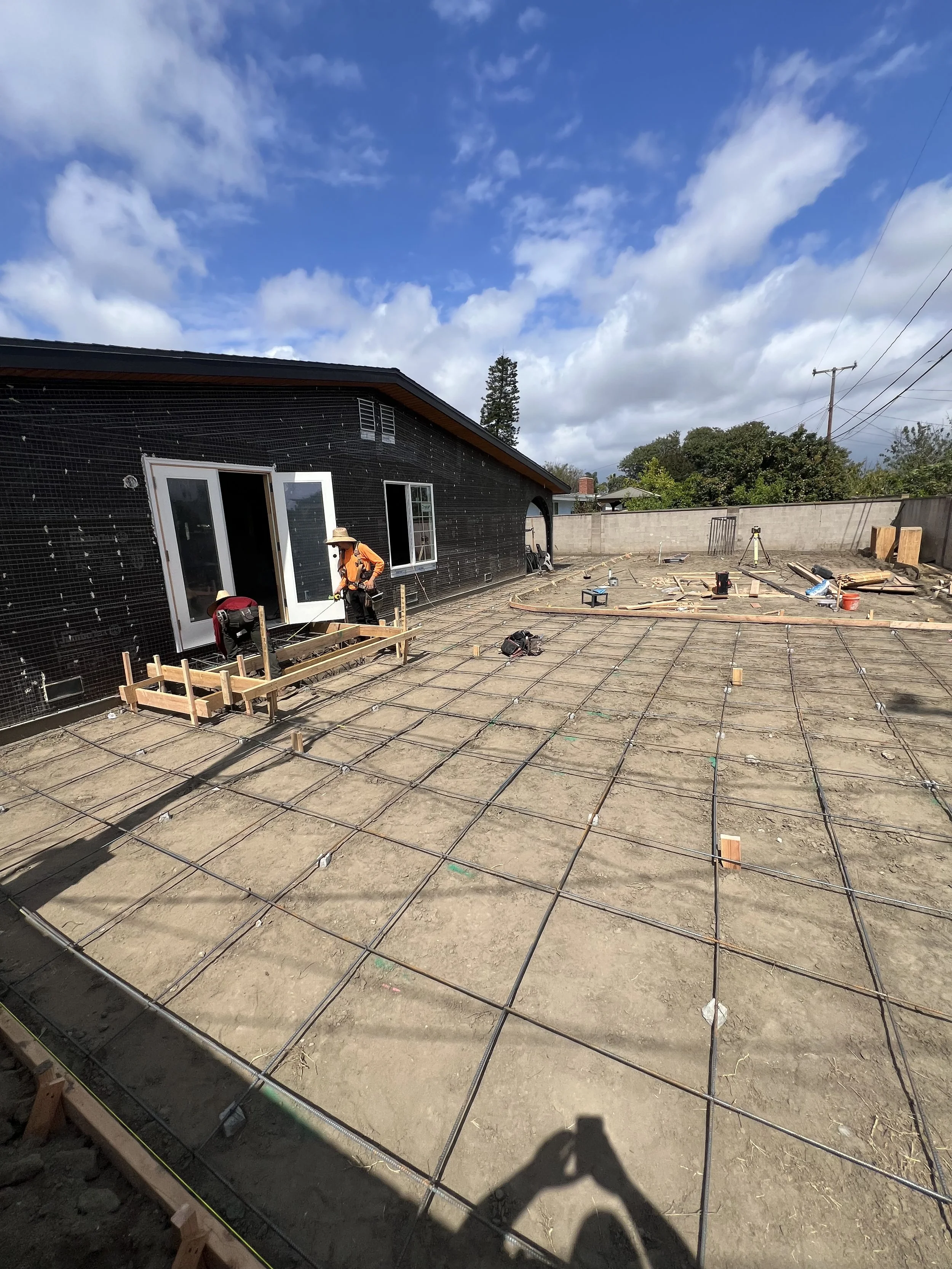 Construction site in process of building a house, with workers preparing the foundation with rebar grid for concrete pour, and a black house exterior with open doors and windows, under a partly cloudy sky.