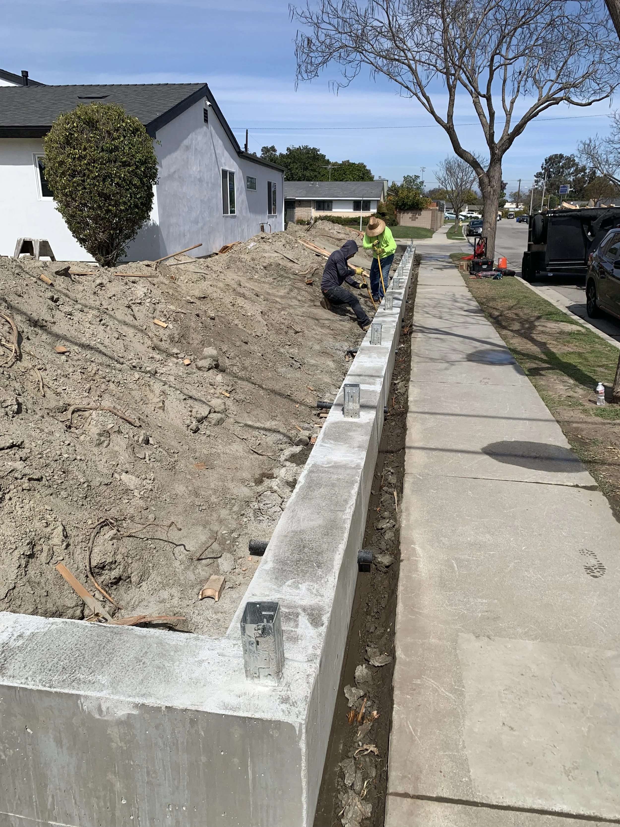 Two construction workers, one wearing a hat and bright yellow shirt, working on a concrete wall next to a sidewalk in a residential neighborhood, with a tree and parked cars in the background.