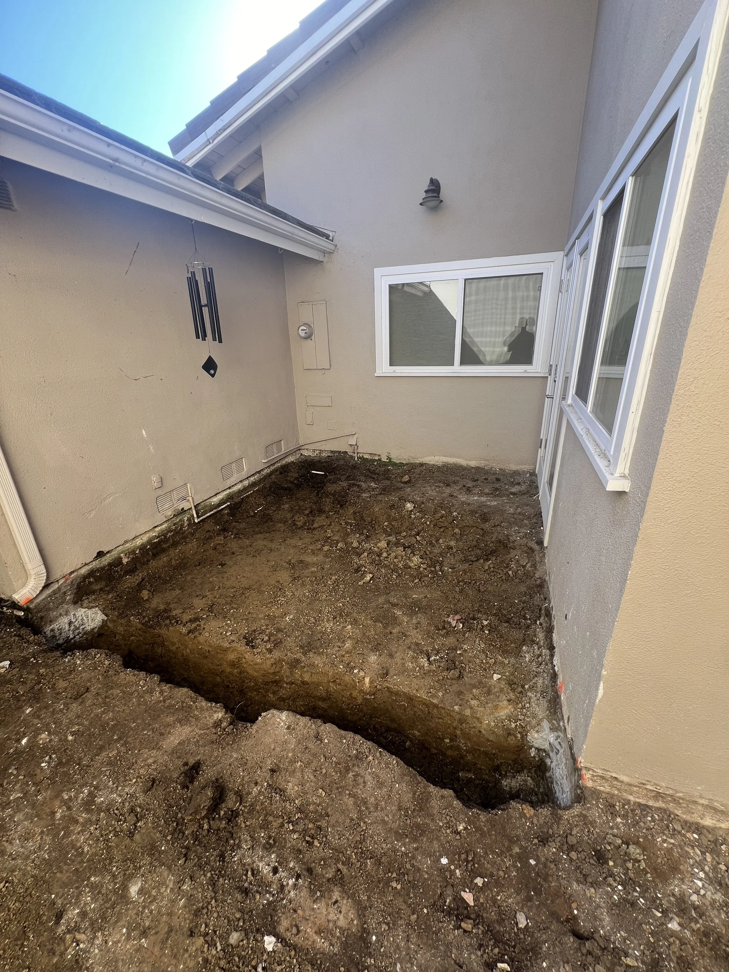 A small backyard patio area partially excavated, with visible soil and dirt, adjacent to the back of a house with beige and gray walls, white-framed windows, and a wind chime hanging from the eaves.