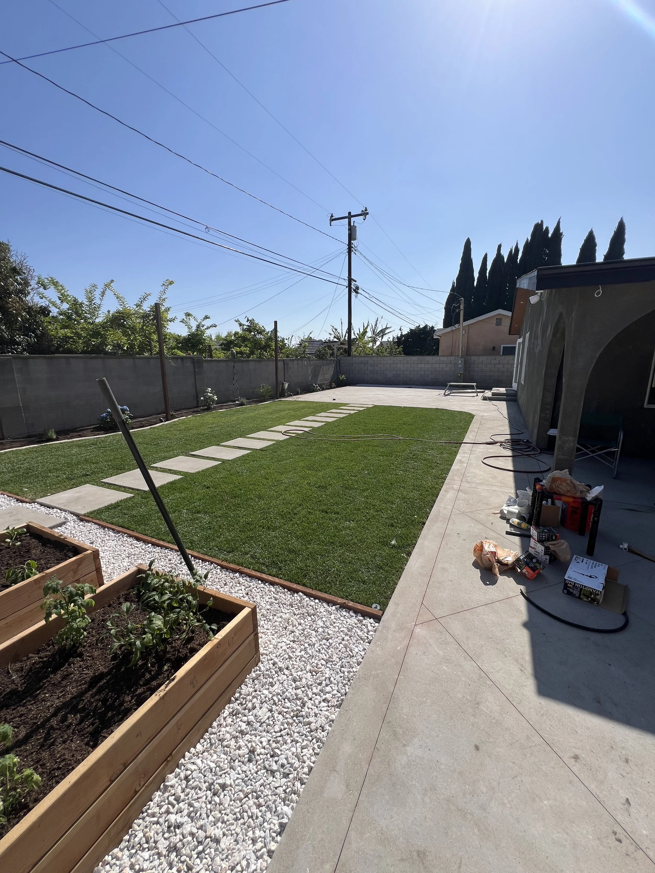 A backyard with a small lawn, stepping stones, a garden bed with plants, and construction tools on the concrete patio under a bright blue sky.