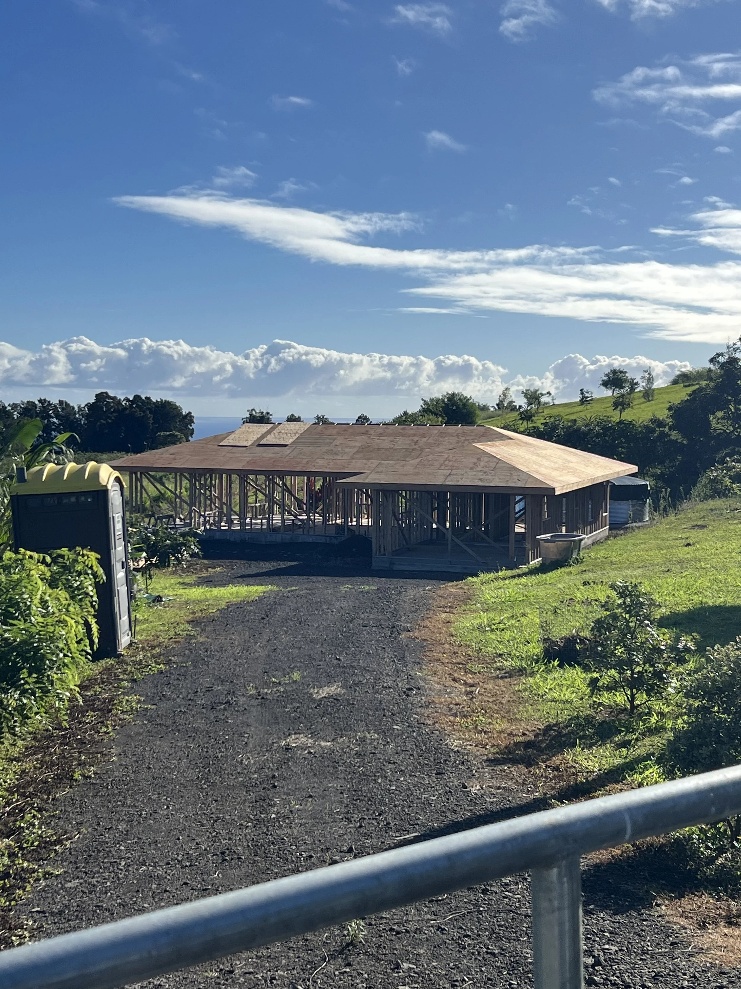 A house under construction with a wooden frame and a partially finished roof, situated on a green hillside with a blue sky and scattered clouds in the background.