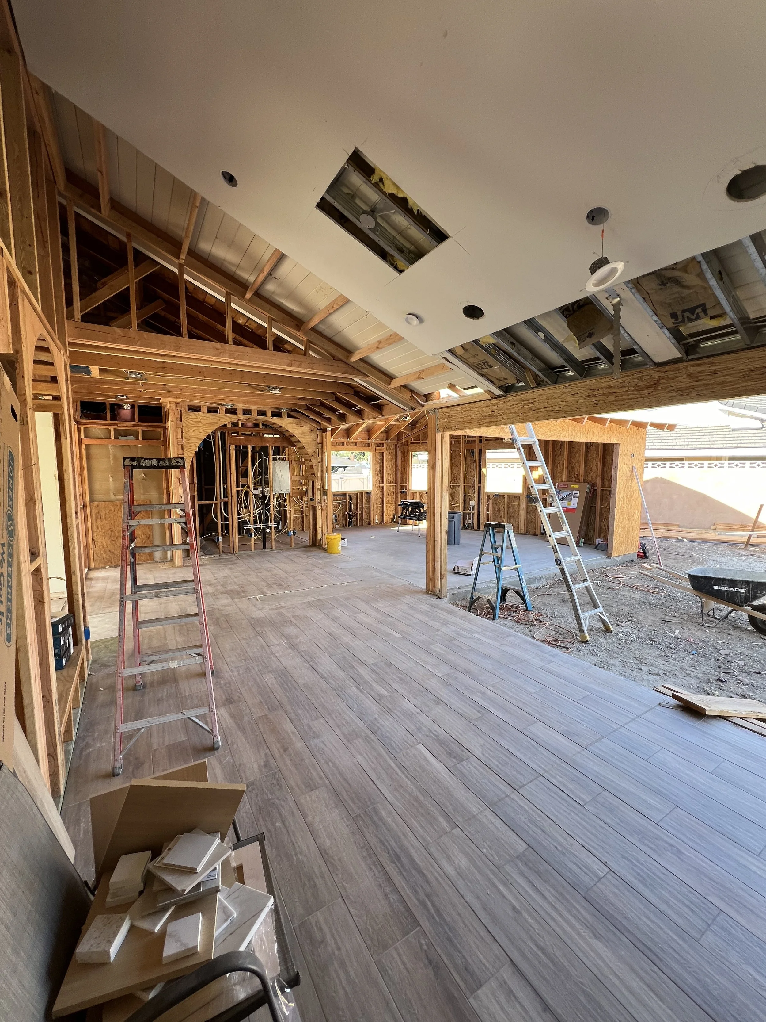 Interior of a house under construction, showing wooden framing, ladders, a partially finished ceiling, construction tools, and flooring.