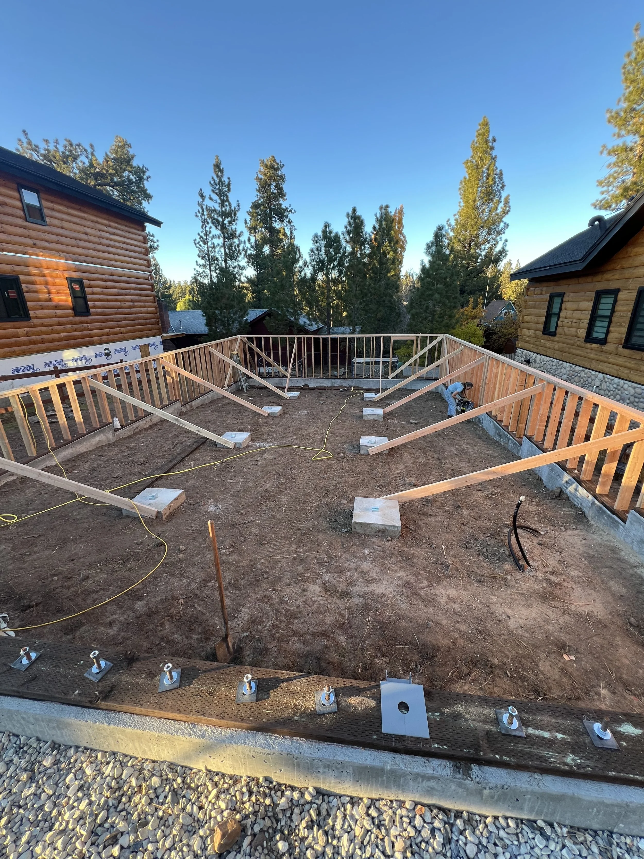 Construction site with wooden framing for a deck or patio, surrounded by two houses and tall trees in the background.