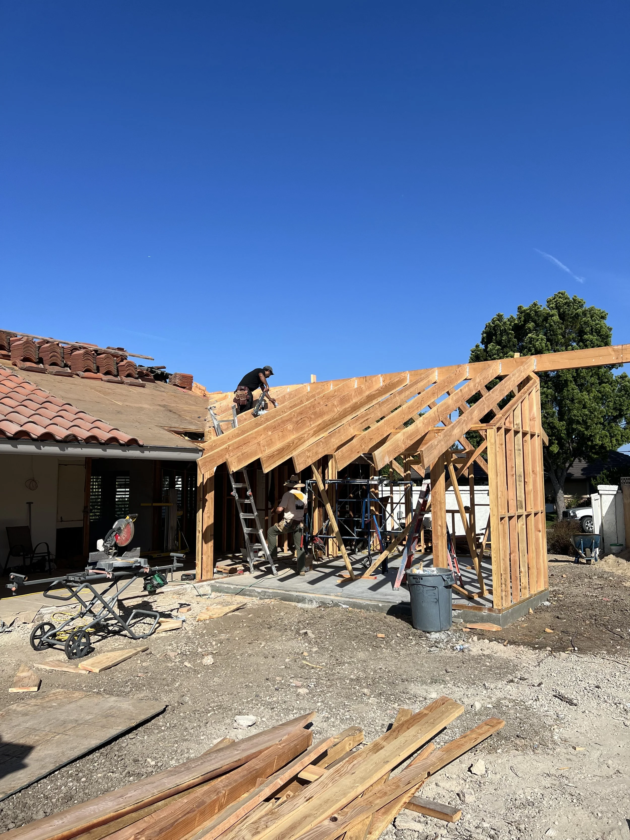 Construction workers building a wooden extension onto a house under a clear blue sky. The framework of the extension is in progress, with some workers on ladders working on the roof structure.