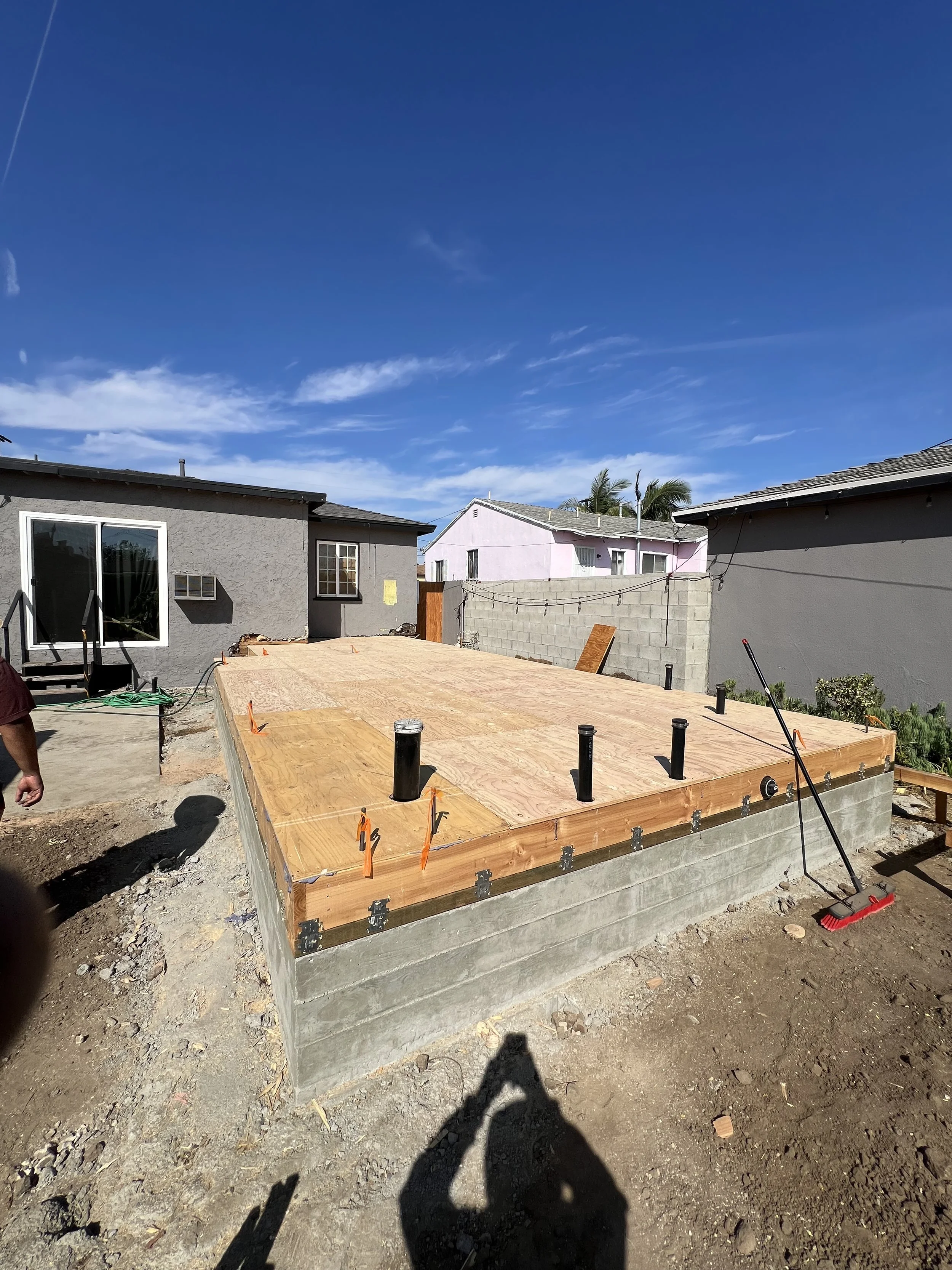 Newly constructed wooden deck with black plumbing pipes, situated on a concrete foundation, in a backyard with neighboring houses and clear blue sky.