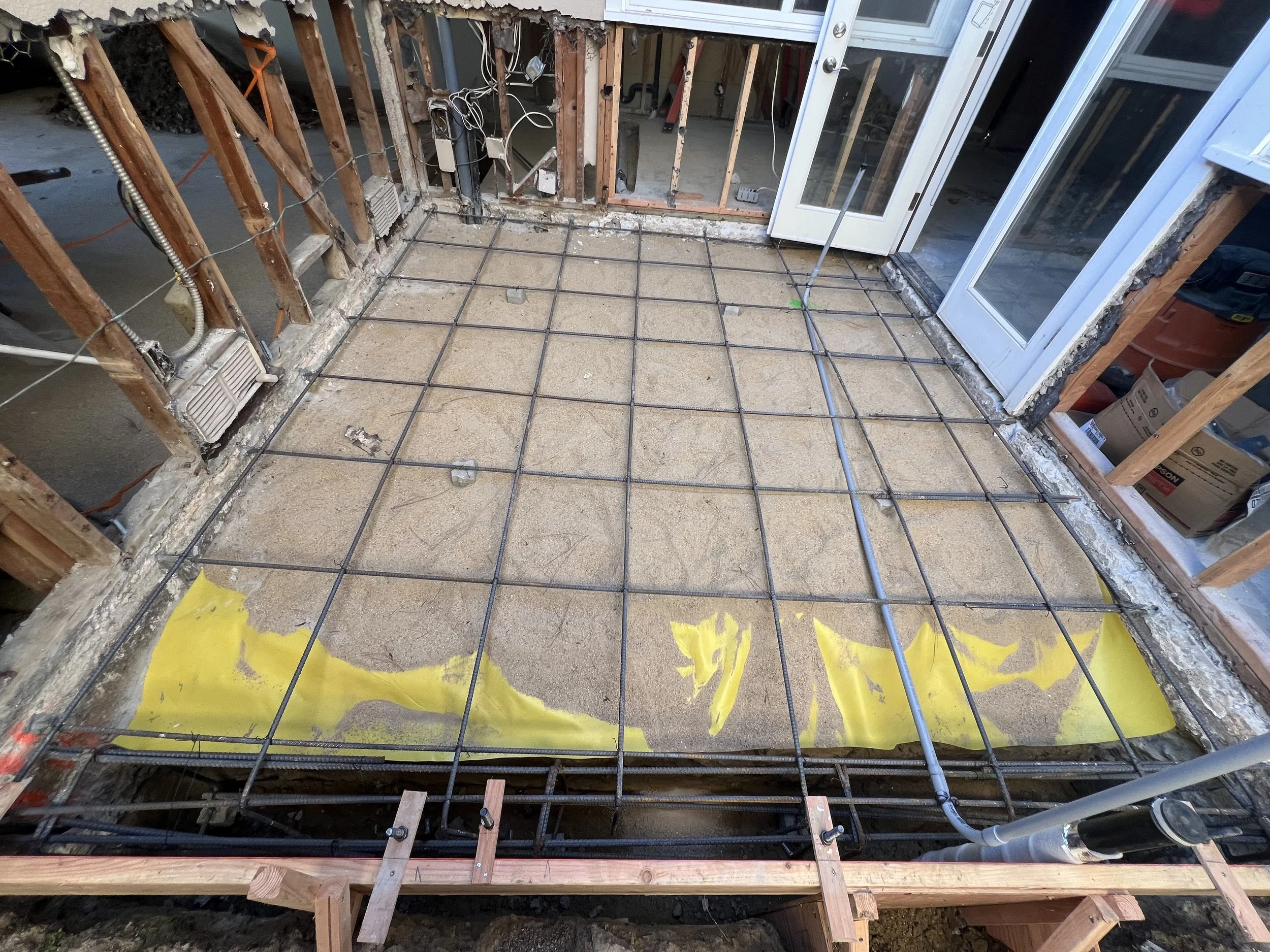 Construction site with a concrete floor prepared with a grid of rebar for pouring a new concrete slab, with a partially yellow waterproofing membrane visible at the edges, and behind glass doors.