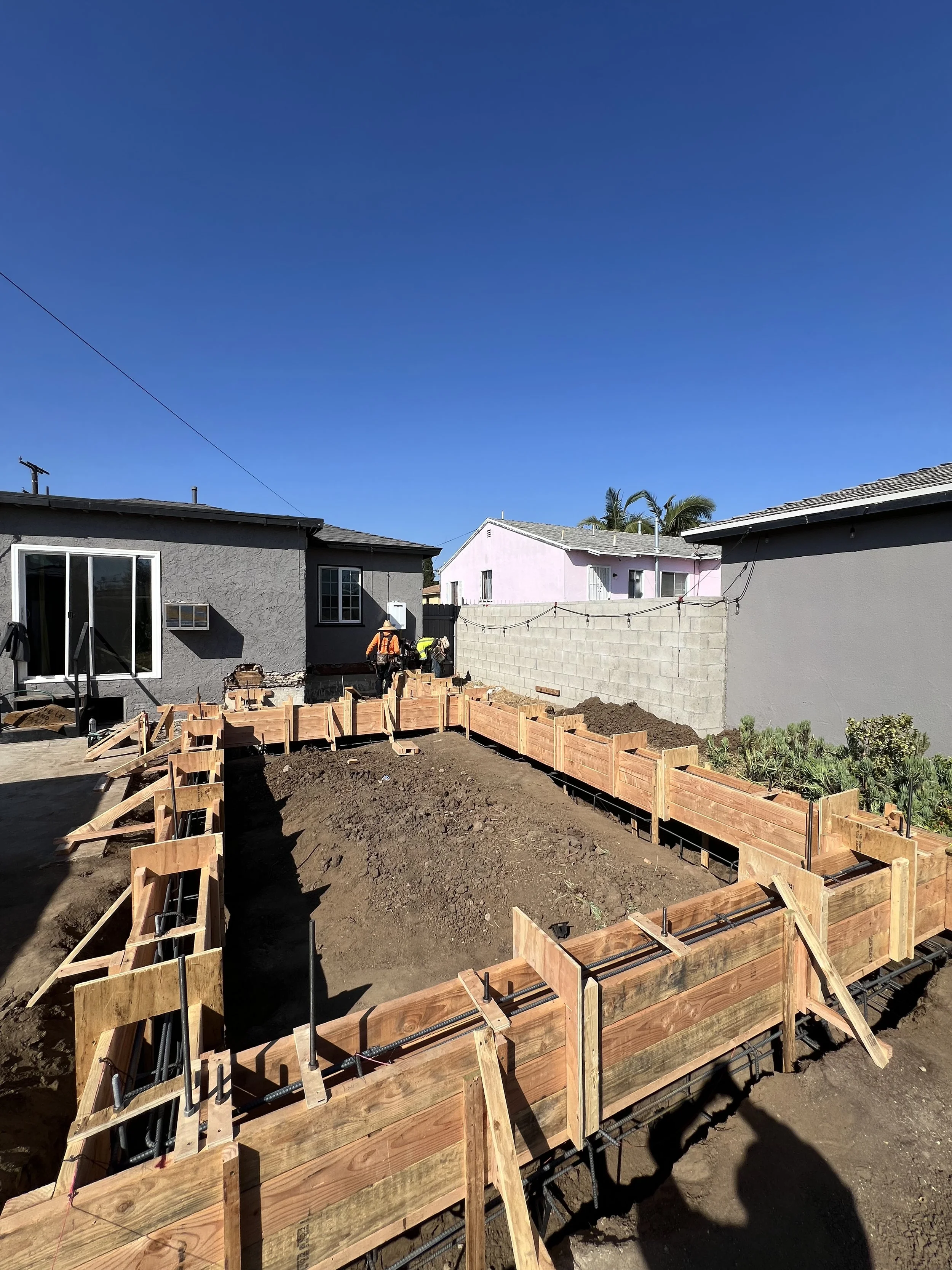 Construction site with wooden formwork for a foundation in a backyard, workers in safety gear, neighboring houses, and clear blue sky.